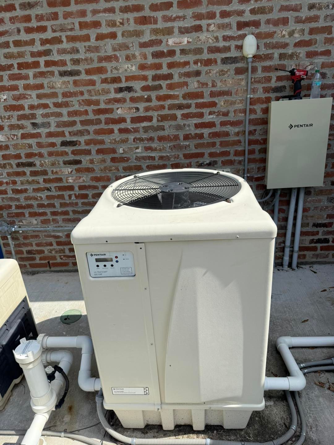 Tan pool heat pump against a brick wall, with white pipes and electrical box.