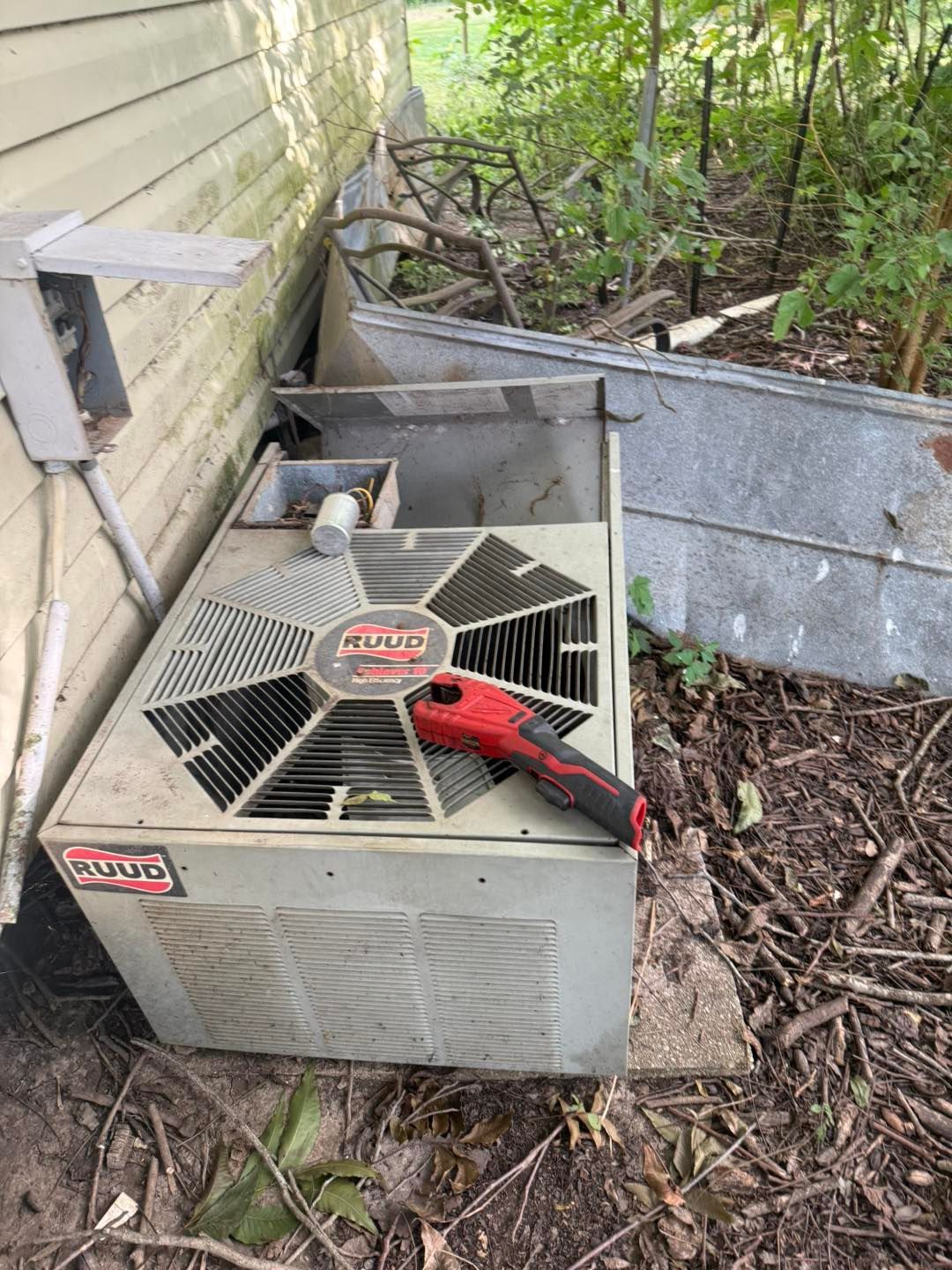 Exterior air conditioning unit with a red tool on top, next to a building and overgrown vegetation.