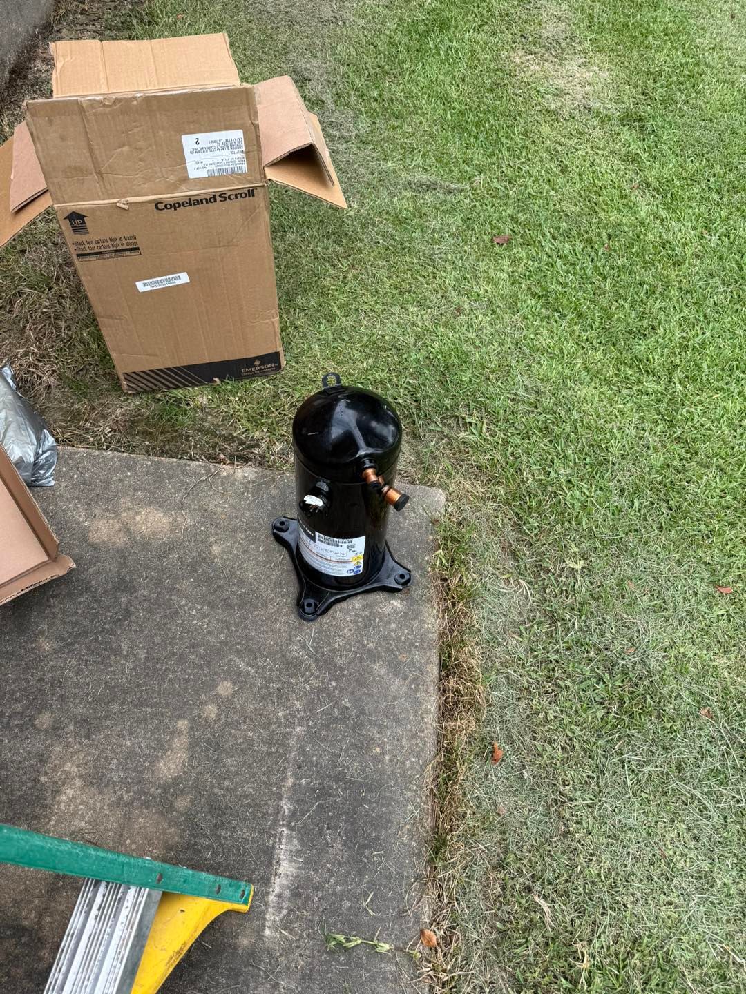 Black air conditioning compressor on concrete next to open cardboard box and green grass.
