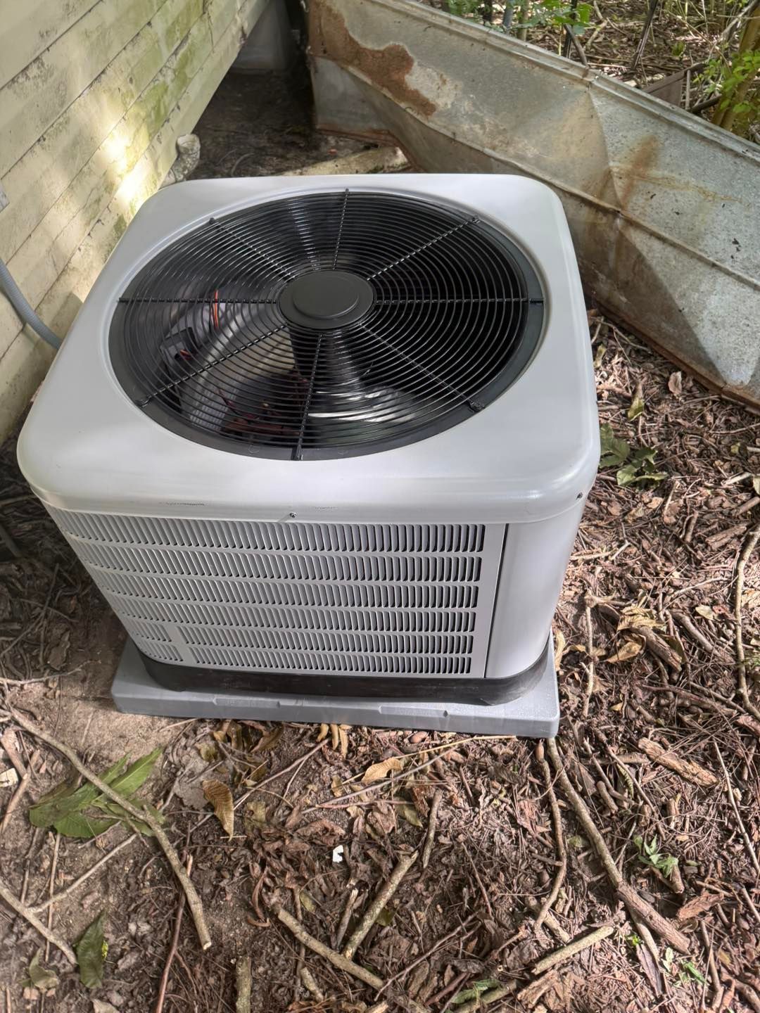 Air conditioning unit, outdoor, gray and black, sitting on a concrete base, near wood and leaves.
