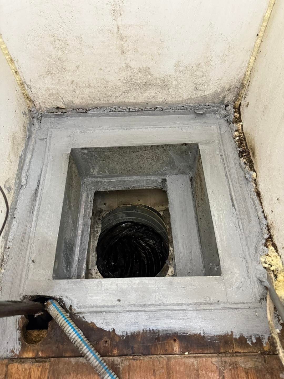 Square opening in the floor with a vent and gray sealant. Wood floor and ceiling visible.