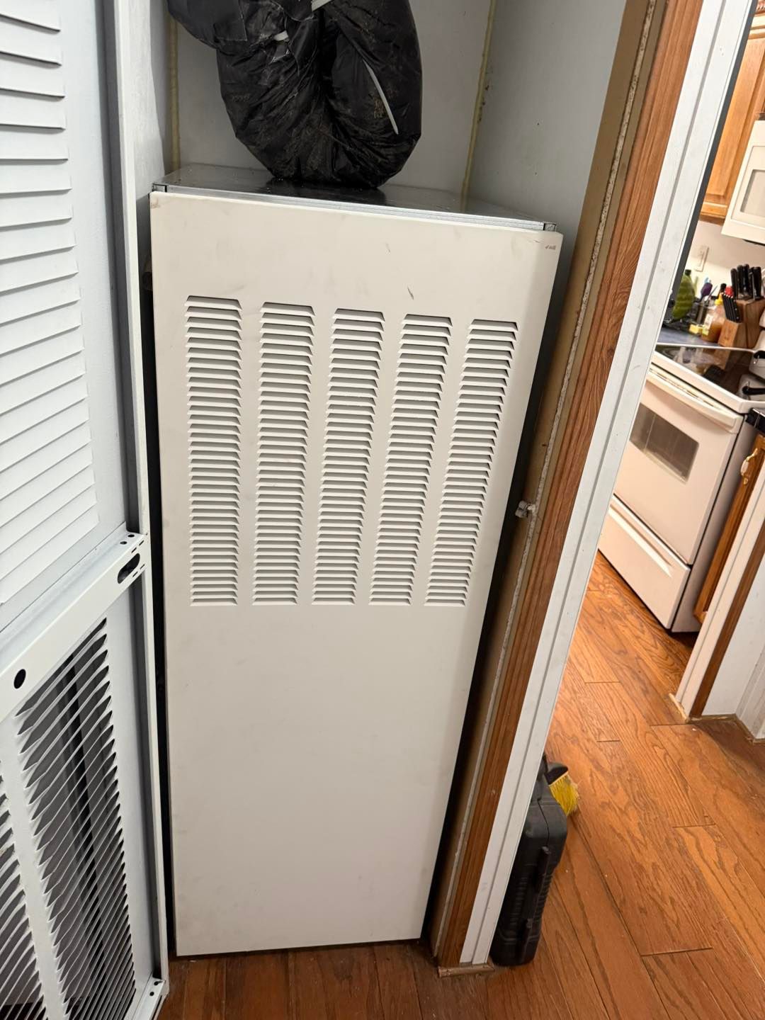 White HVAC unit with vent slits, located in a narrow space with an open door and air vent.