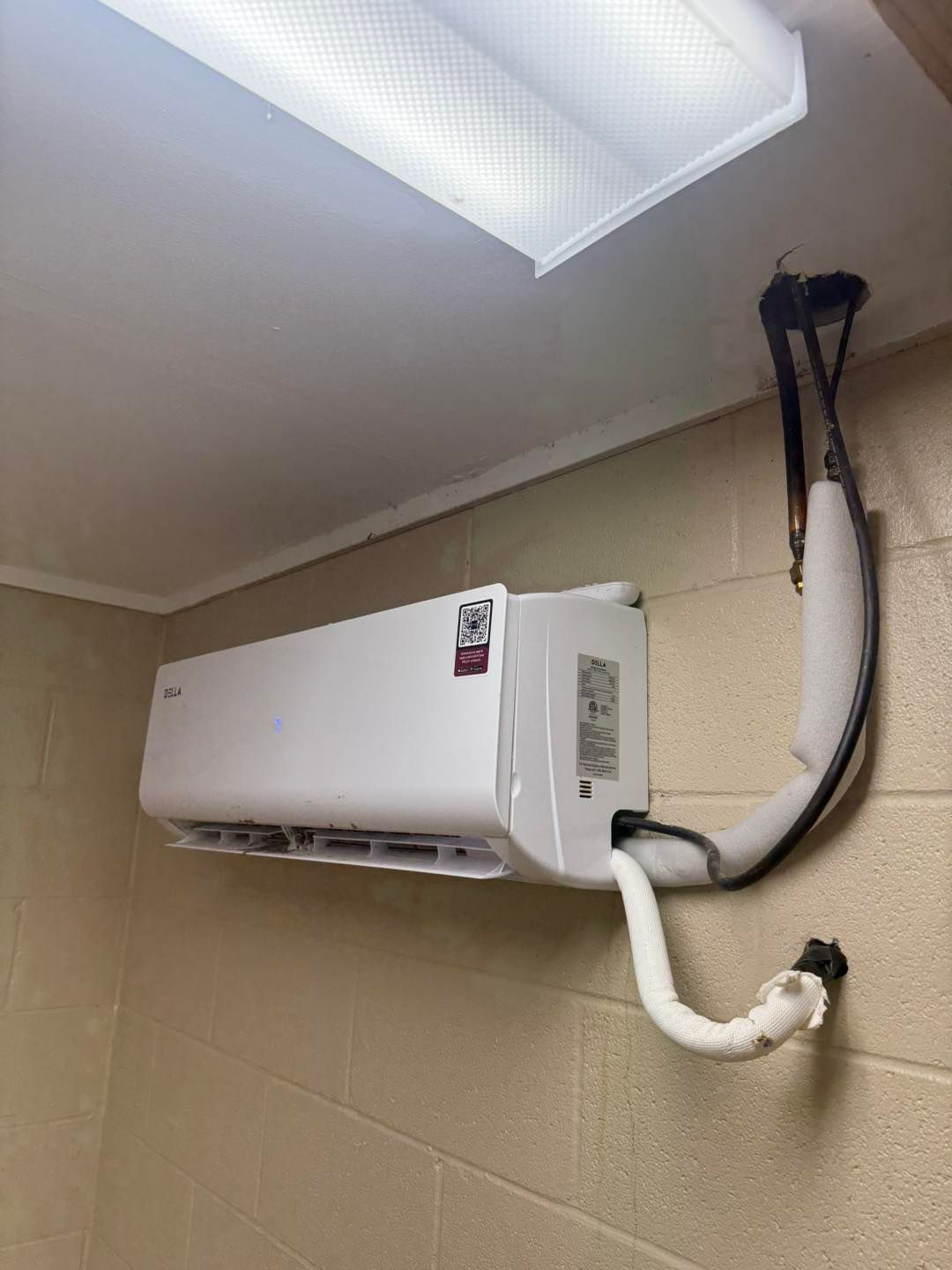 White wall-mounted air conditioner unit installed on a light beige brick wall. Flexible pipe curves up and out.