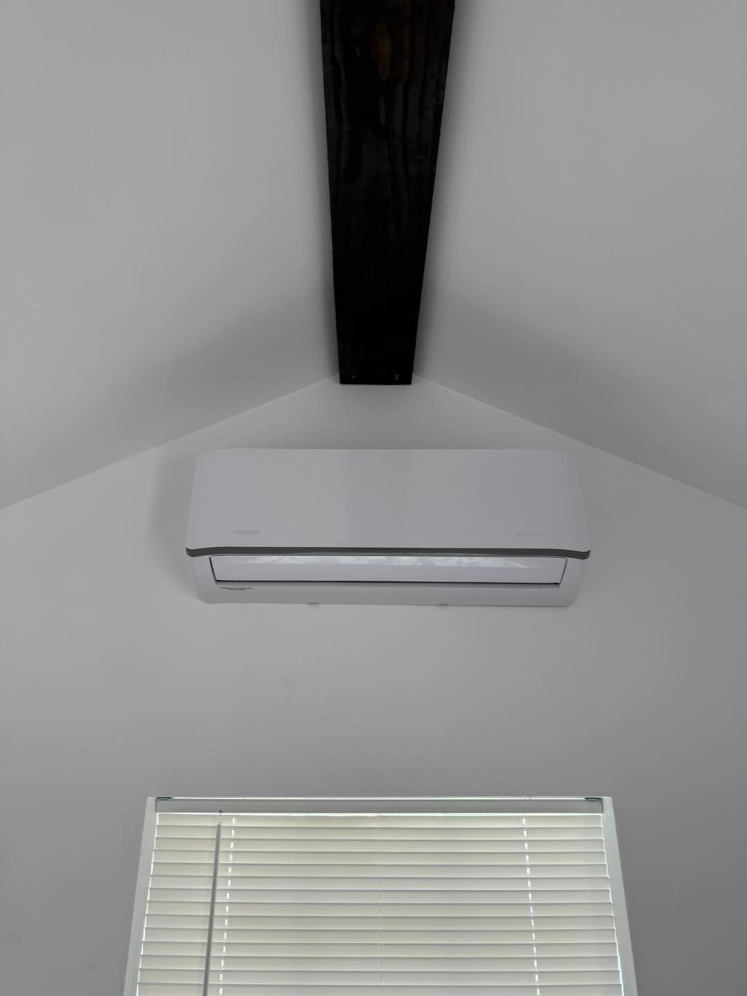 White air conditioner on a white wall below a black wooden beam, with a window below.