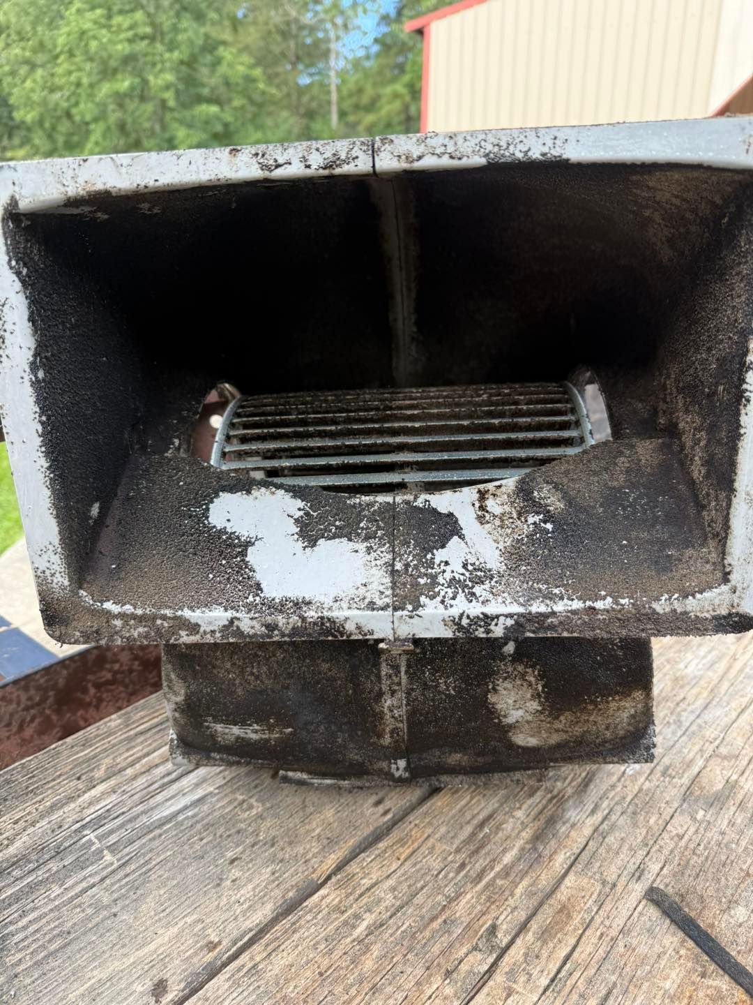 A rectangular, gray metal vent with a dirty fan inside, set on a wooden surface.