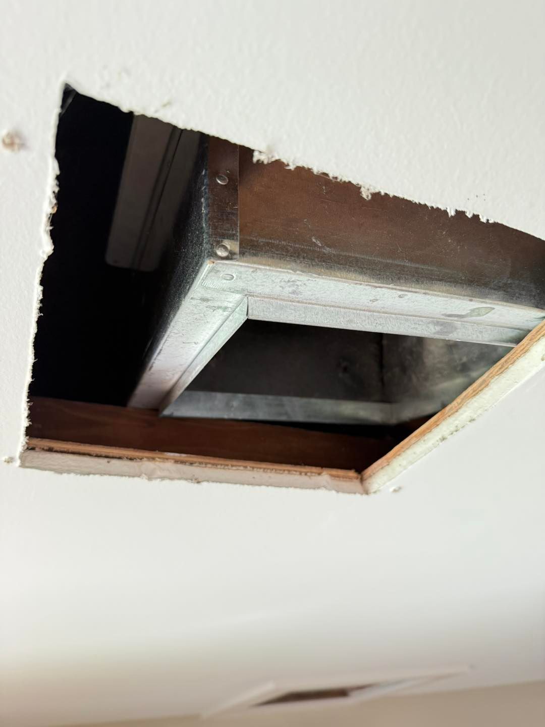 Ceiling opening revealing a metal duct and wood frame, in a white ceiling.