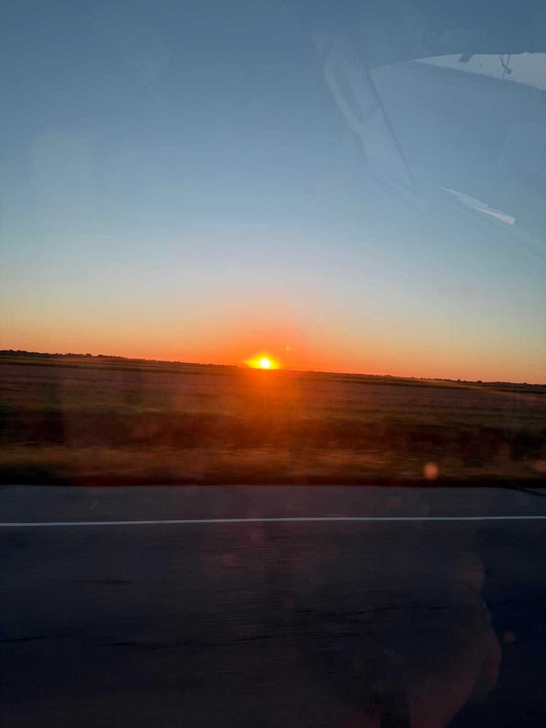 Sunset over a field, viewed from a vehicle; orange sun, blue sky, highway.