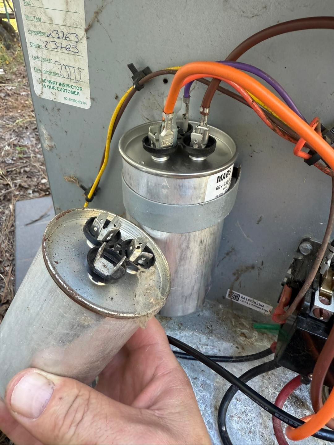 A hand holding a cylindrical capacitor next to one connected to an air conditioning unit.
