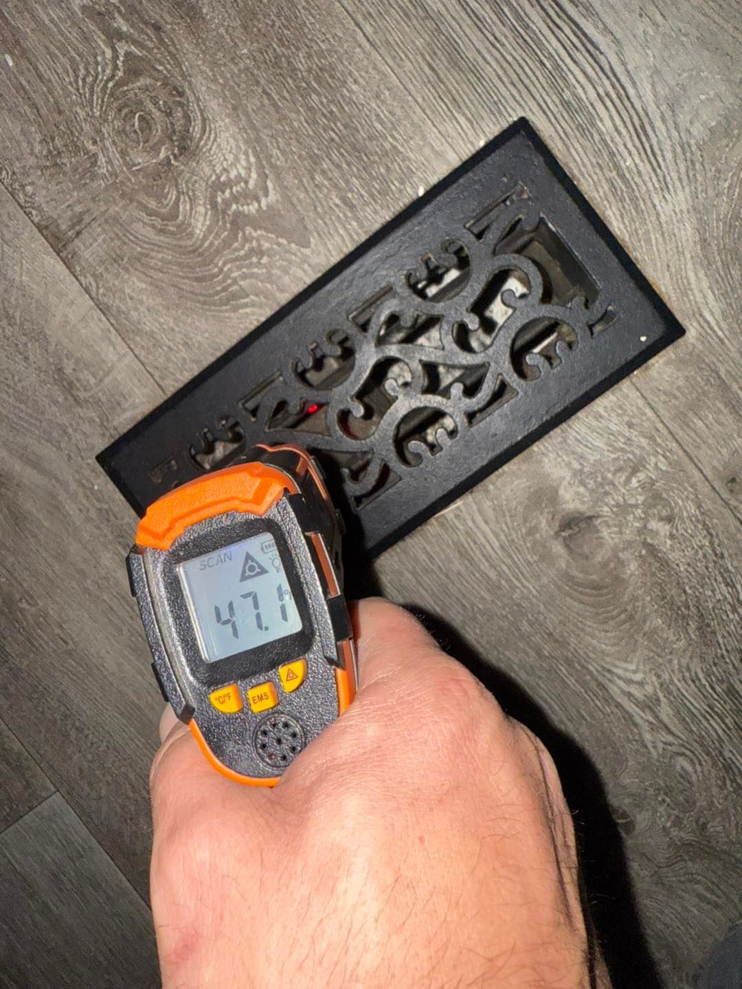 A hand holding a temperature gun measures 47 degrees Fahrenheit at a floor vent.