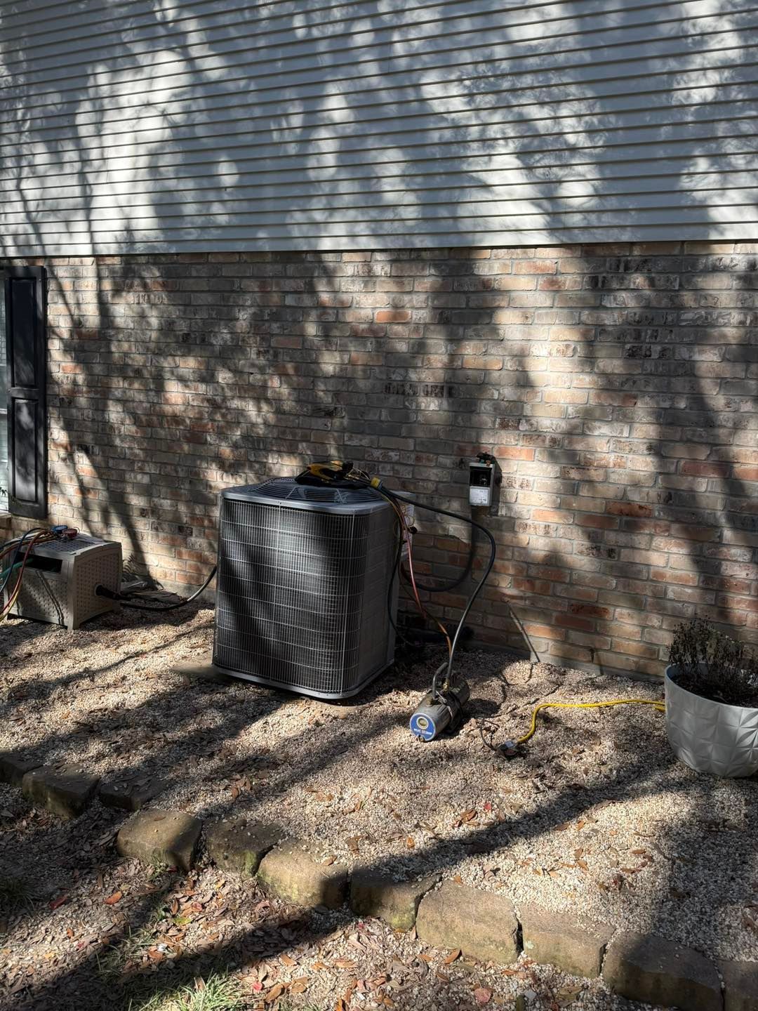 An air conditioning unit sits outside against a brick wall, connected to an electrical box and drain.