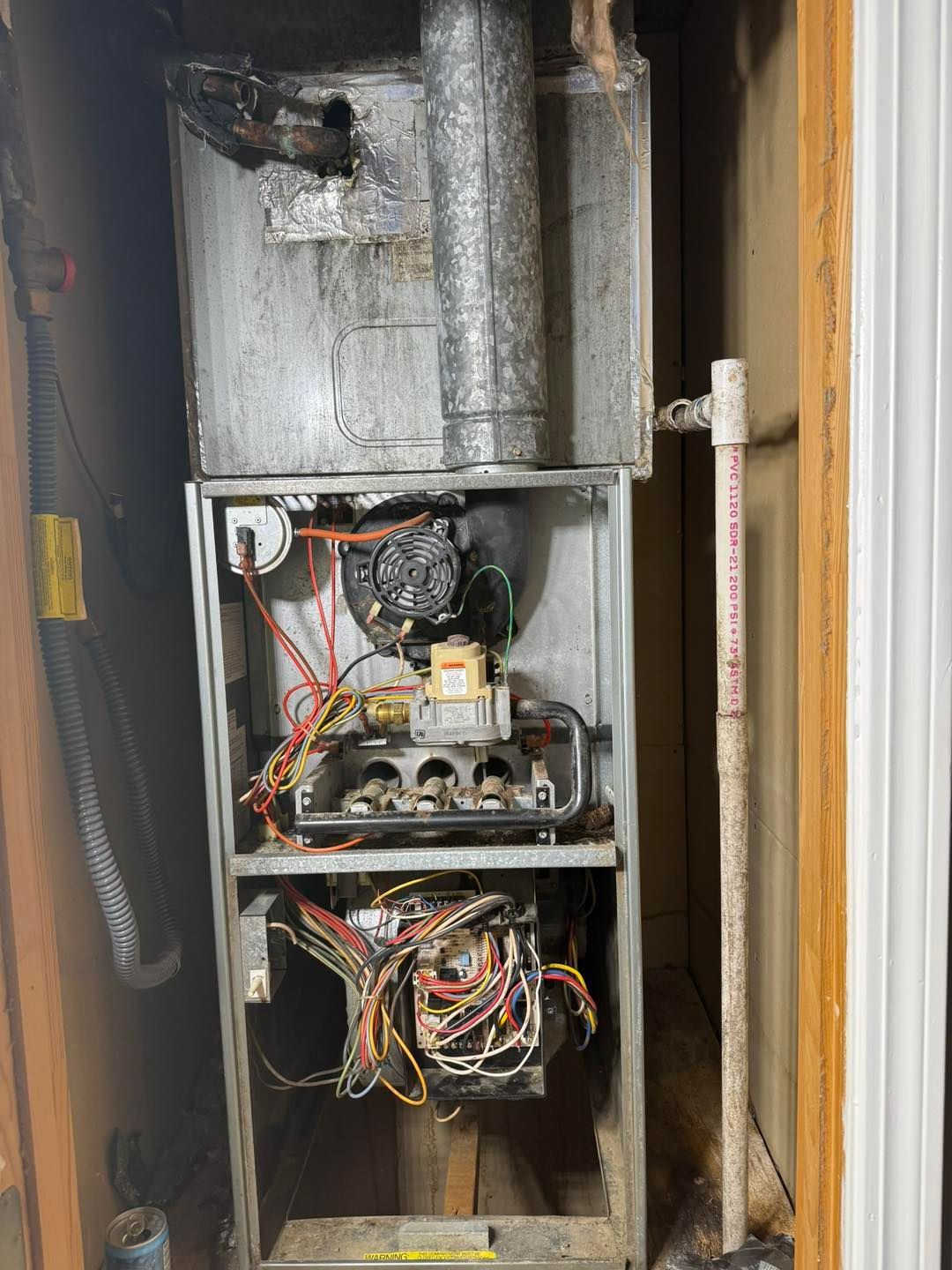Furnace in a utility closet with exposed components and vent pipe. Wiring visible.