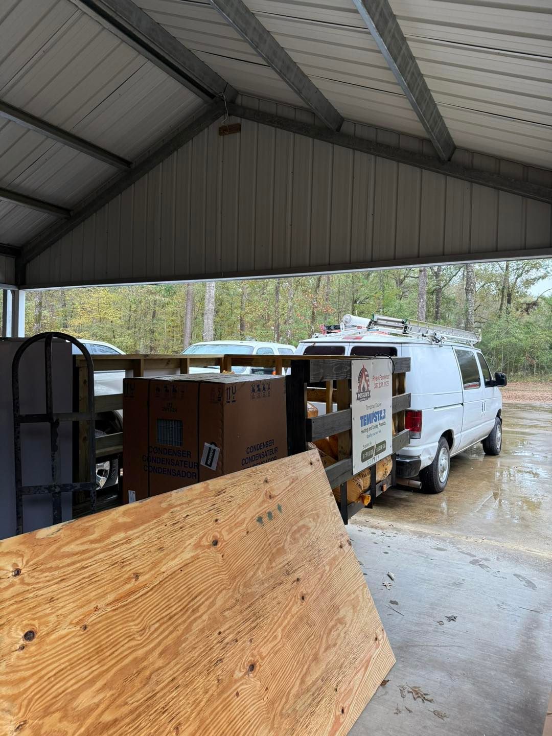A white van parked under a metal-roofed structure with construction materials visible, possibly an open-air carport.