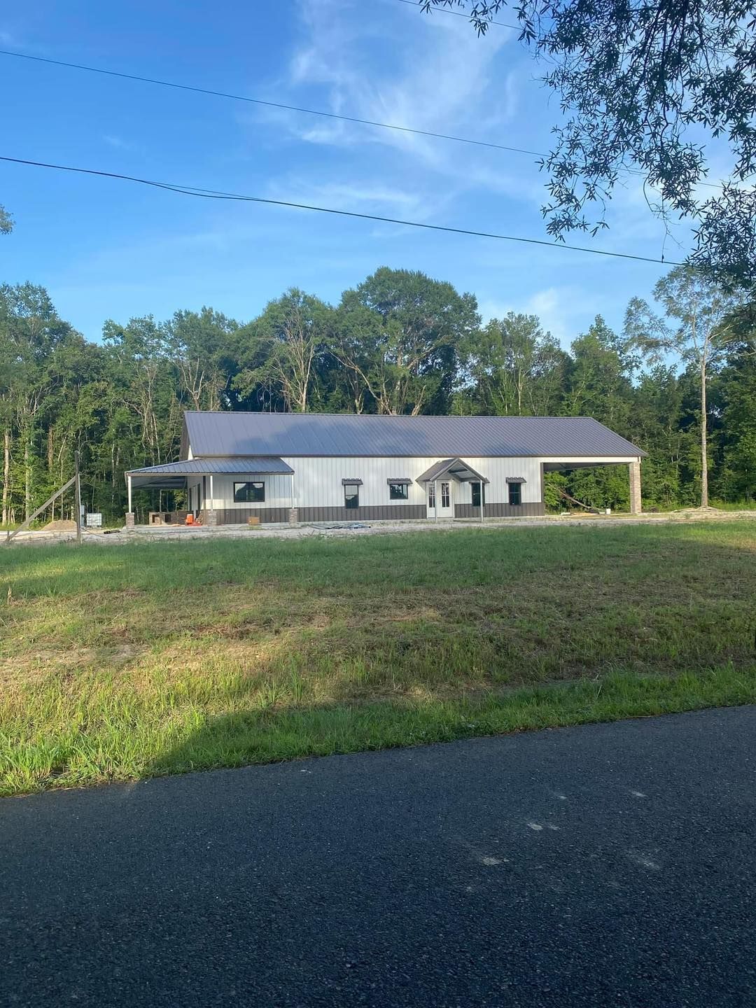 New, white building with gray accents, a dark roof, and covered porch on a grassy lot with trees in the background.