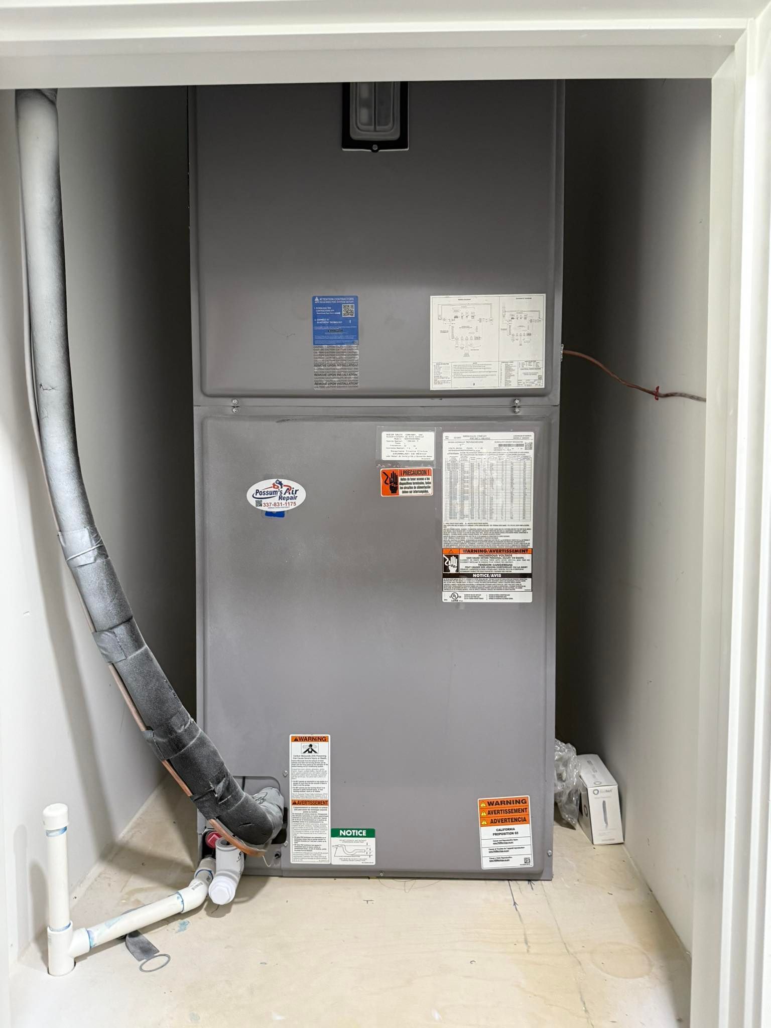 Large gray HVAC unit in a white-walled utility closet with insulated ducting, pipes, and warning labels.