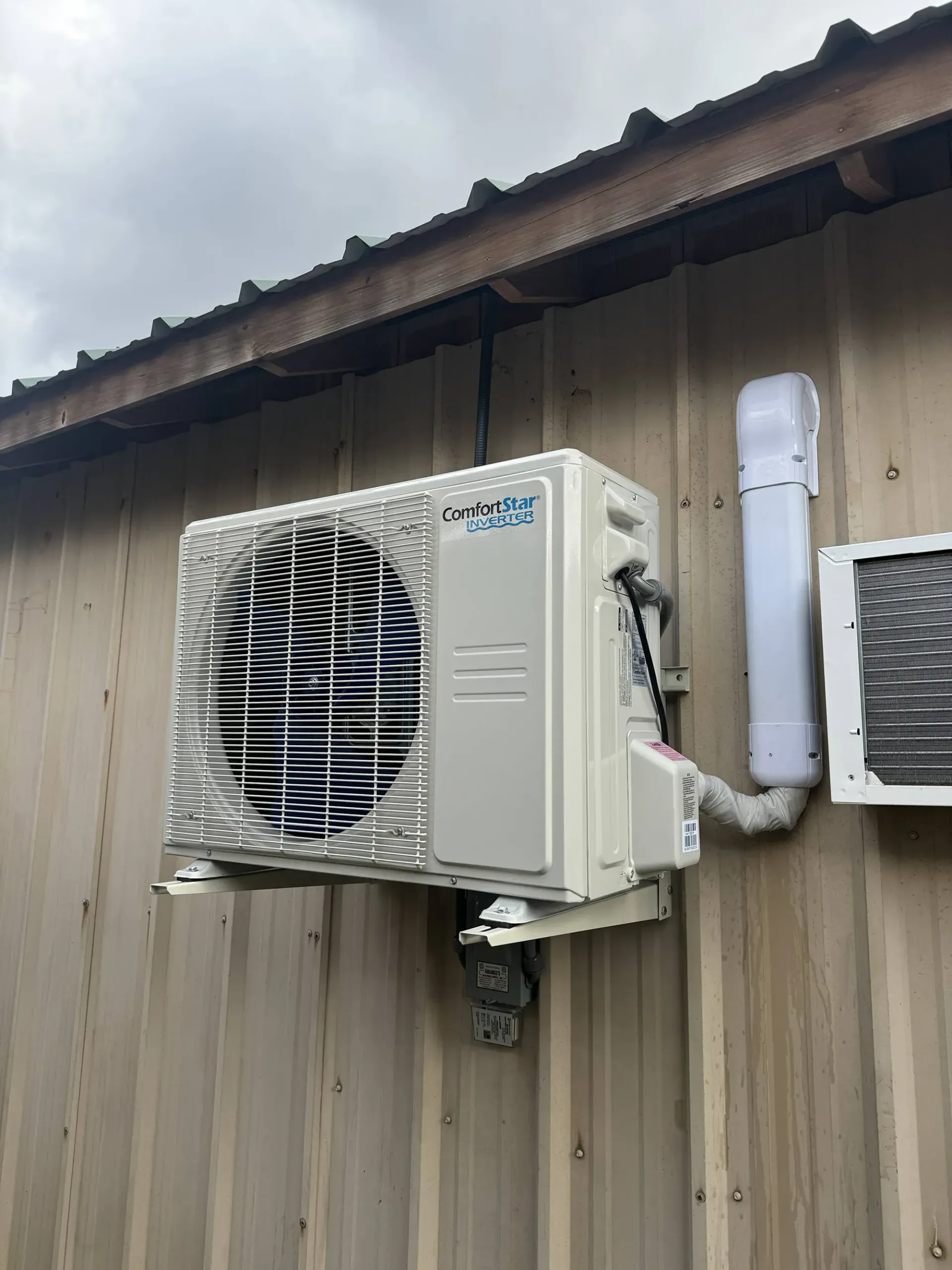 Outdoor air conditioning unit mounted on a wooden wall beside a window and vent.