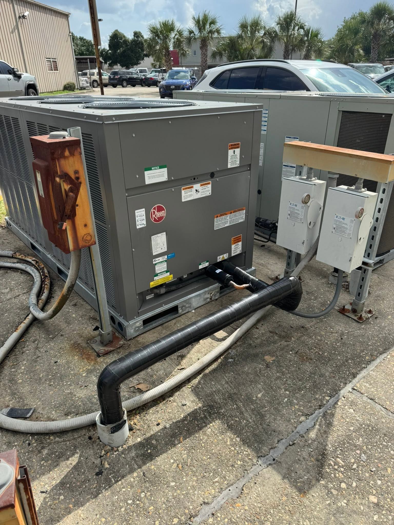 Gray electrical utility box and conduit pipes on gravel beside a parking lot, with cars in the background