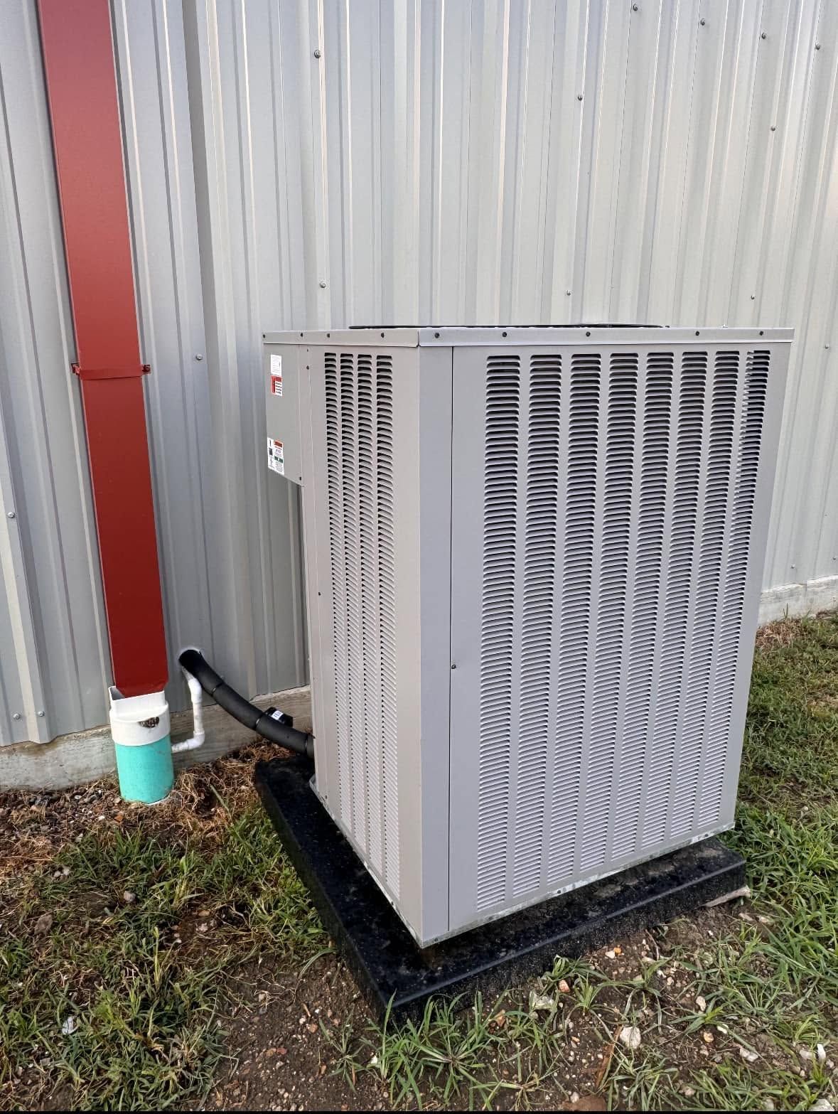 White outdoor HVAC unit beside a red downspout and corrugated metal wall on grass.
