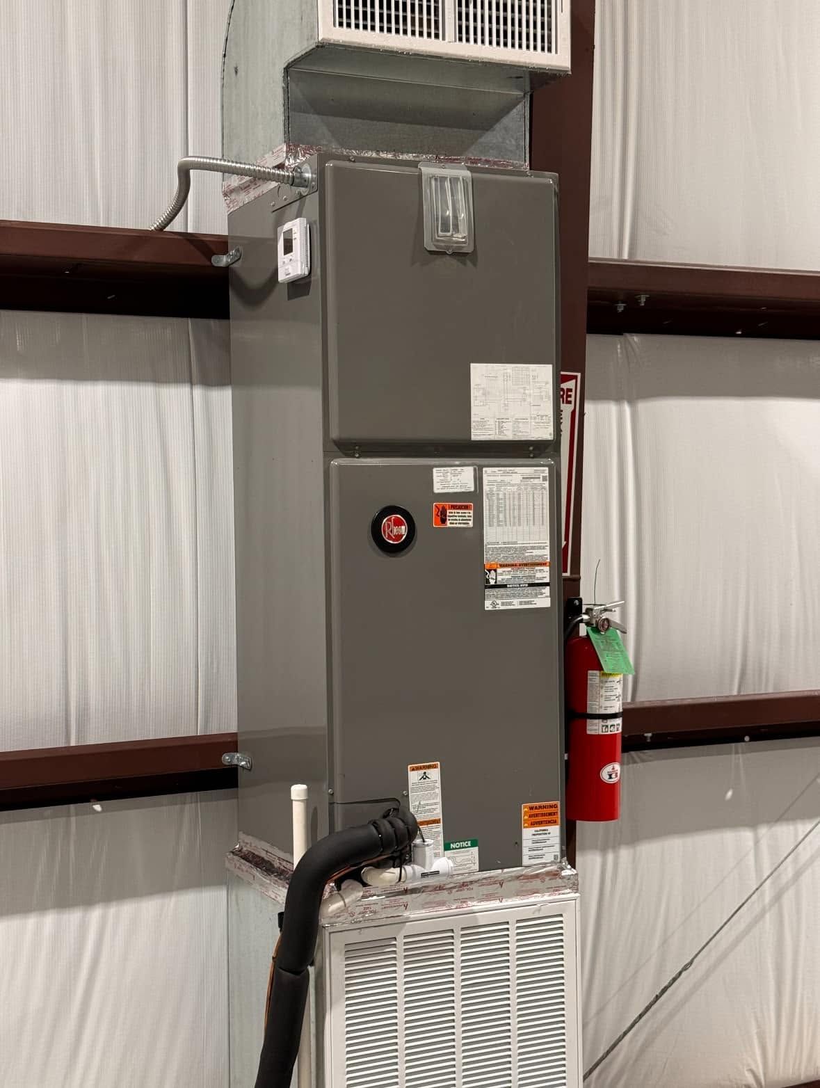 Gray HVAC unit in a metal-walled utility room, with a red fire extinguisher beside it.