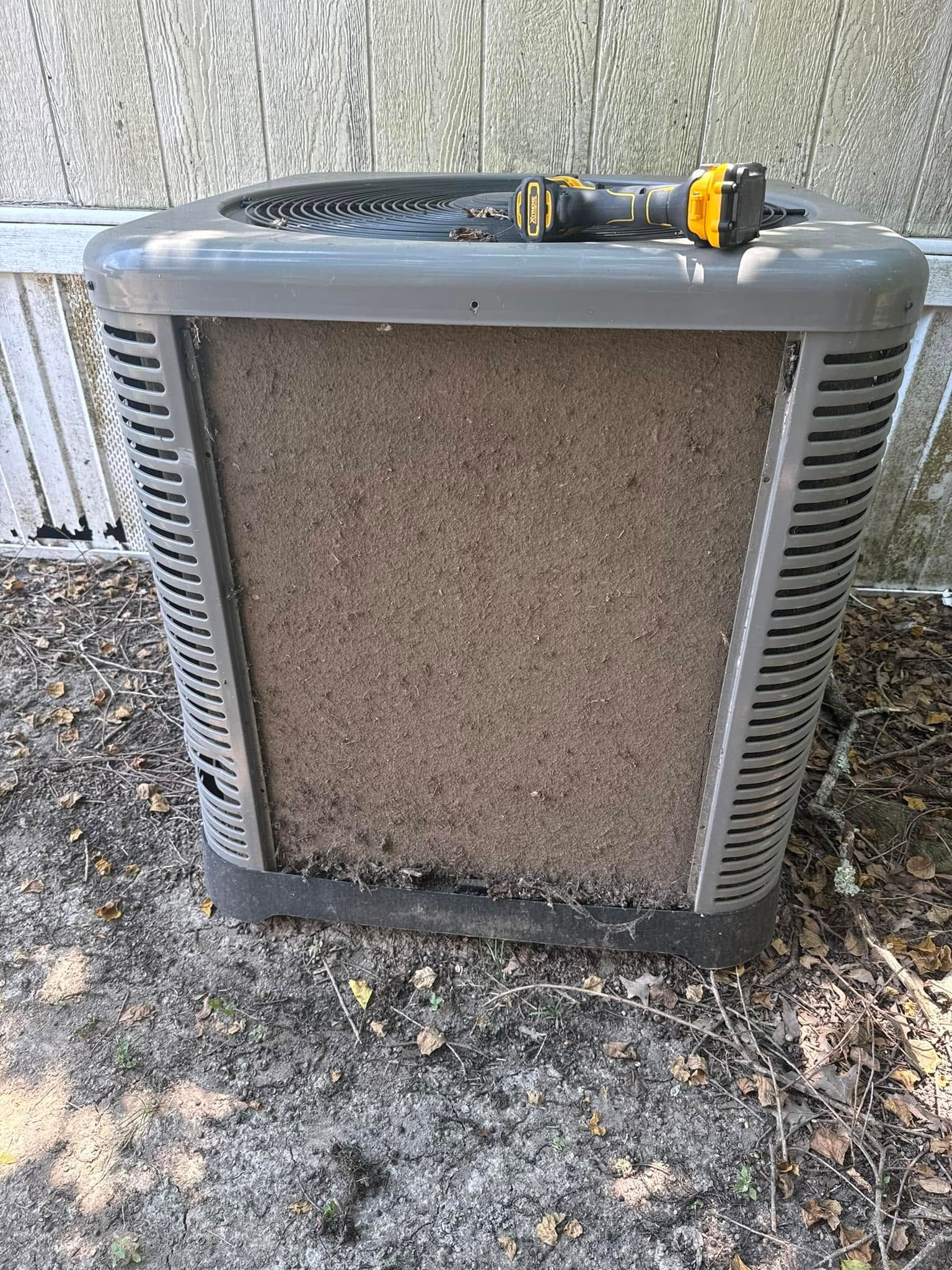 Dirty outdoor HVAC unit with a yellow hose tool resting on top.