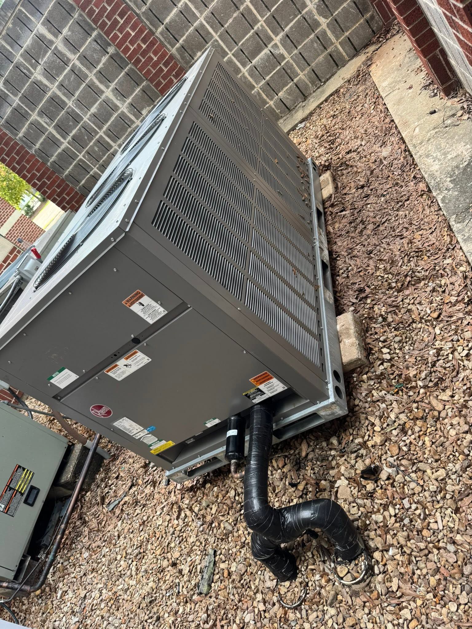 Outdoor HVAC unit beside a brick wall, with black pipes and gravel ground around it