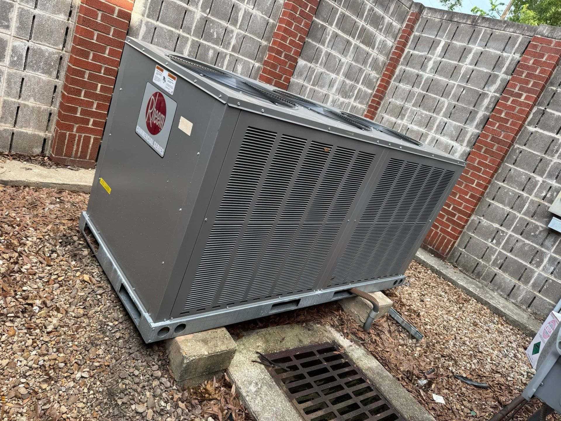 Gray HVAC unit beside brick wall and drain grate in a gravel yard.