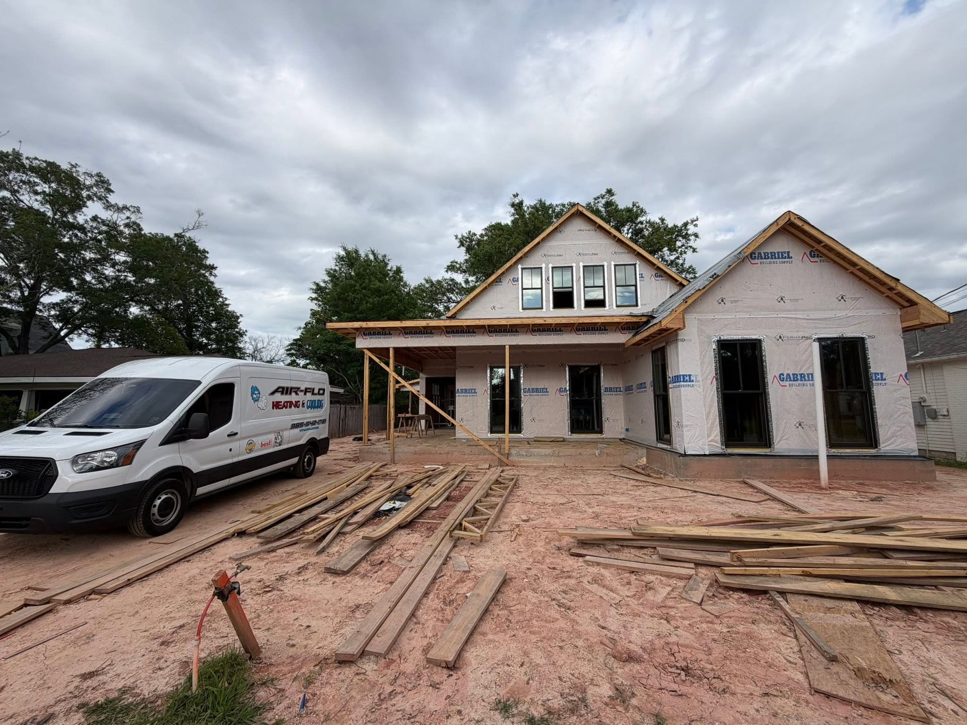 House under construction with a white work van, lumber in foreground, and cloudy sky above