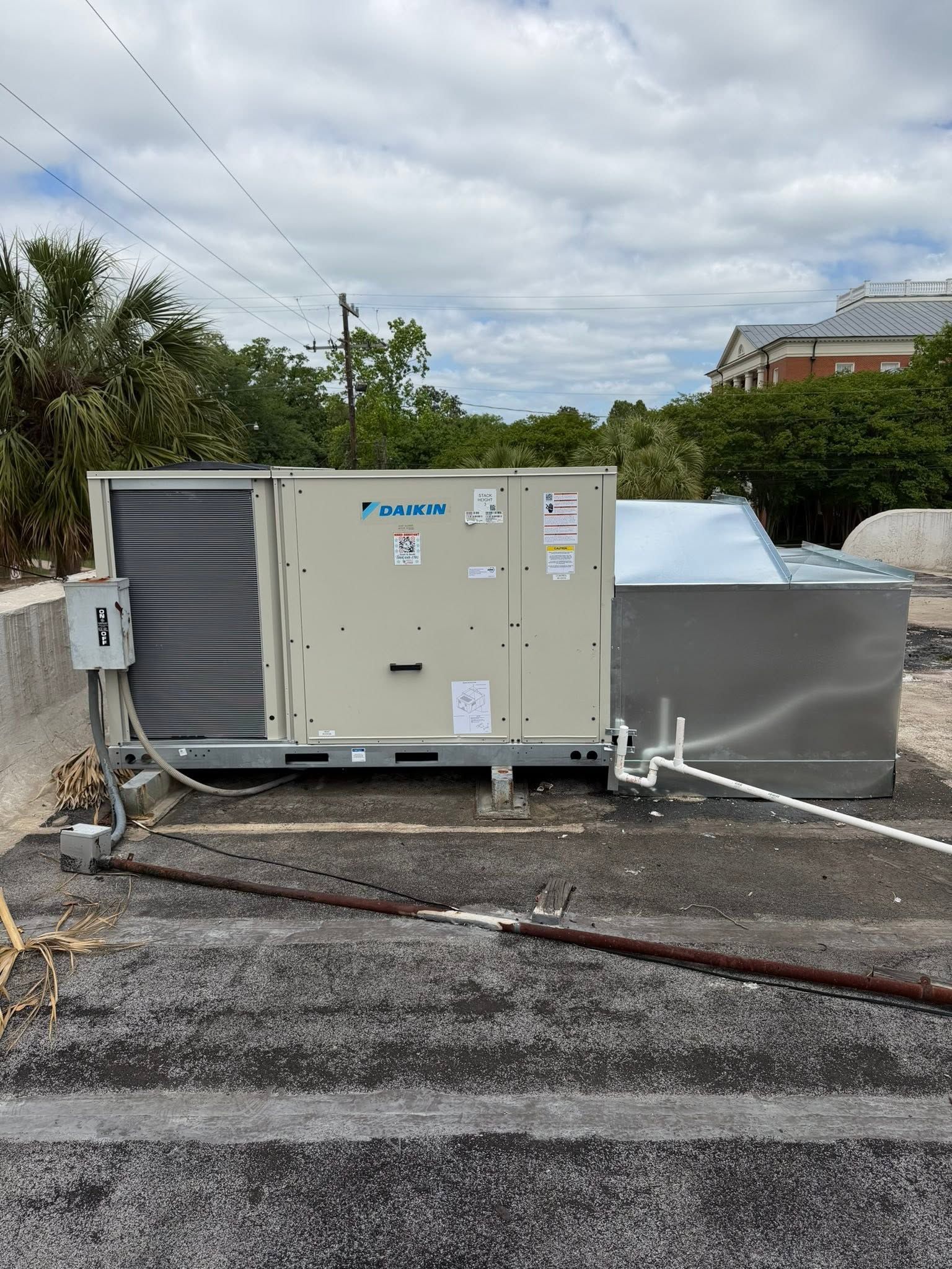 Rooftop HVAC units and ductwork beside gravel and shrubs under a cloudy sky