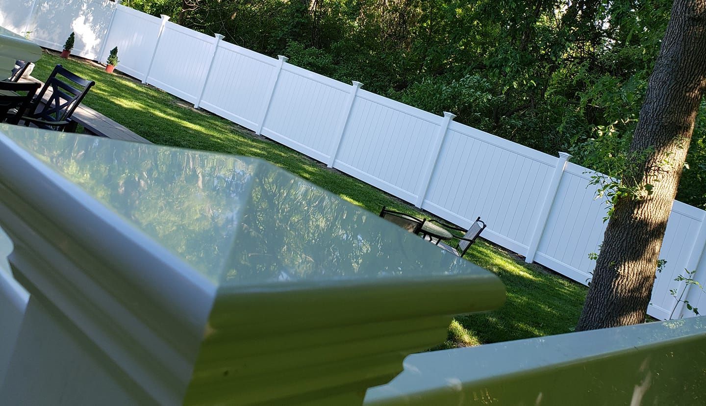 Tilted view of a backyard with a white fence, green lawn, patio table, and trees in bright sunlight