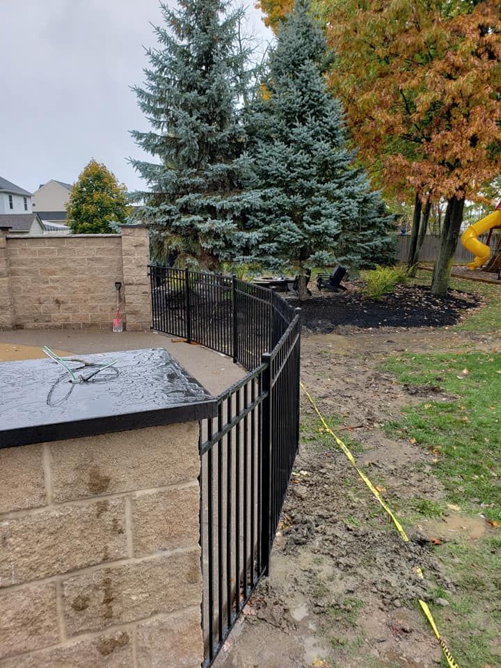 Black metal fence beside a tan stone wall, with evergreen trees and a yard in the background.