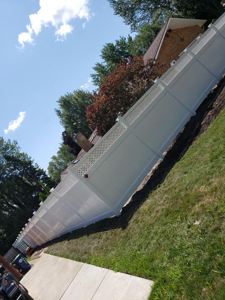 White fence along a sunny sidewalk beside a brick house and trees.