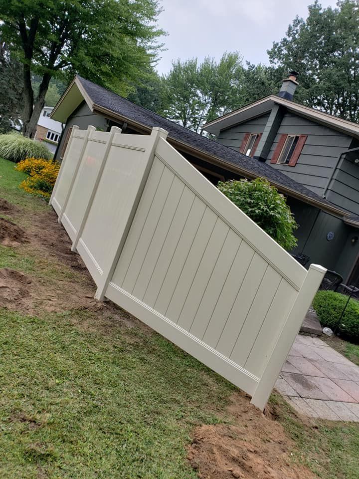 Tilted beige fence panels beside a house in a sloped yard with dirt and grass.