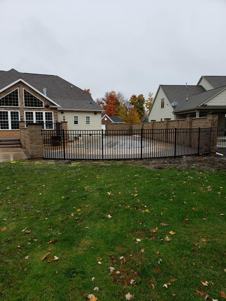 Suburban backyard with a black metal fence, green lawn, and houses under a gray sky.