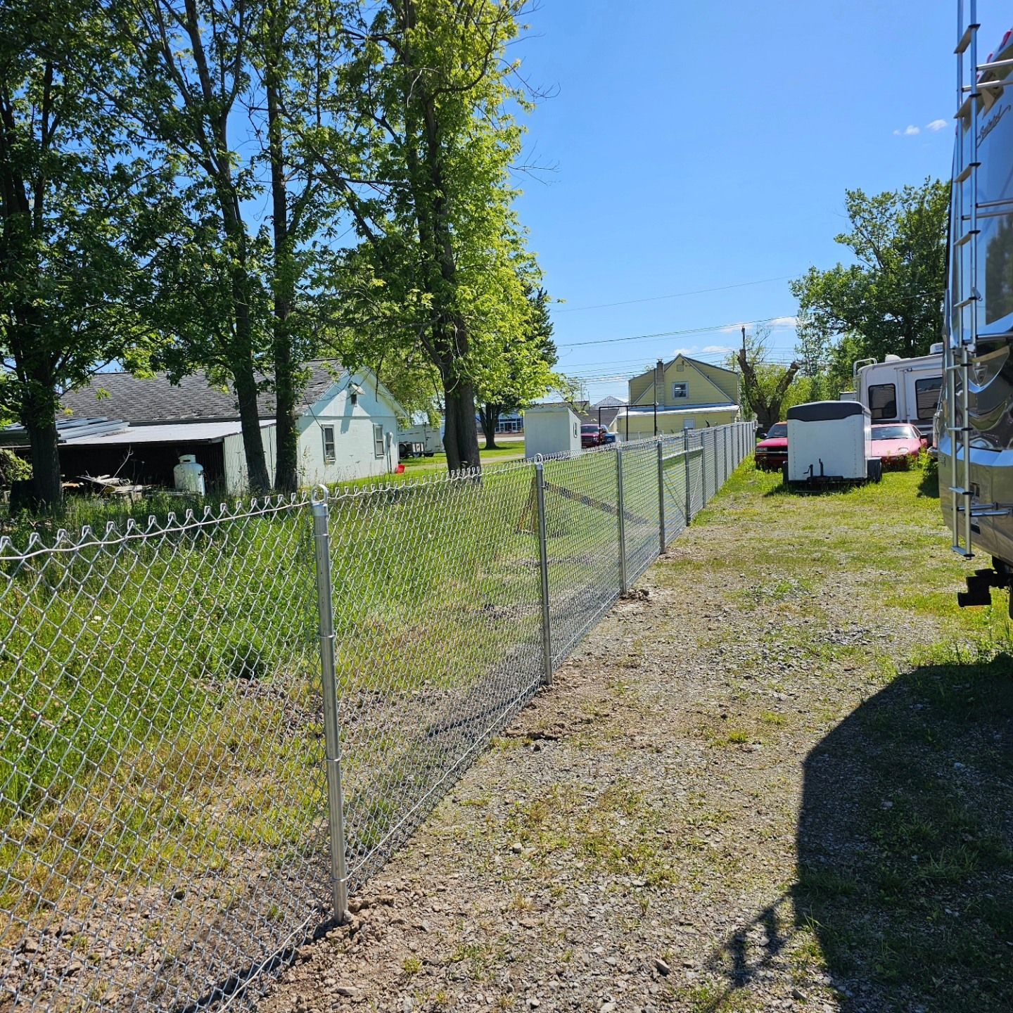 Chain-link fence beside a grassy driveway with trees, small houses, and a white trailer under a blue sky