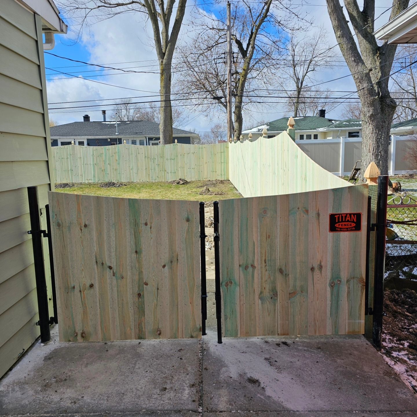 Wooden driveway gate and privacy fence with a small red warning sign, seen from a concrete walkway