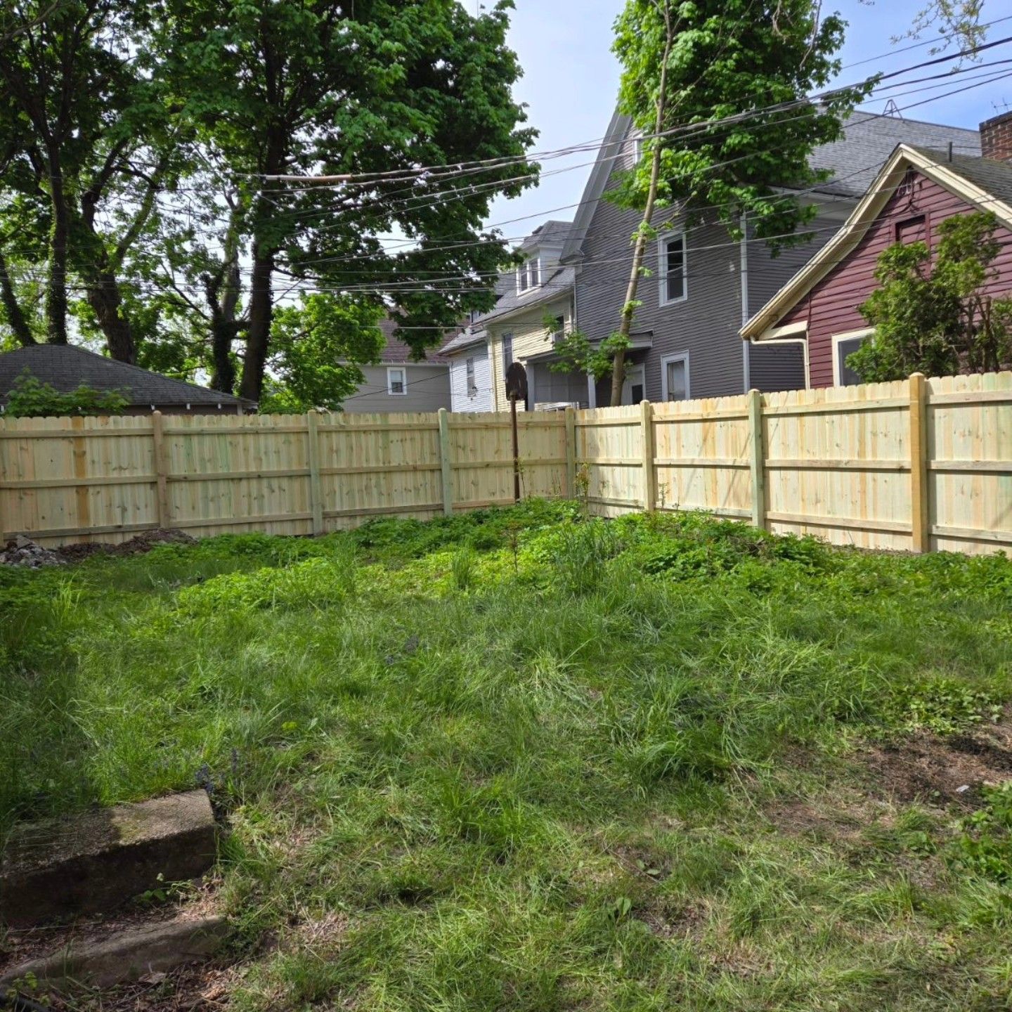 Backyard with grassy yard, light wood fence, trees, and neighboring houses under a partly cloudy sky