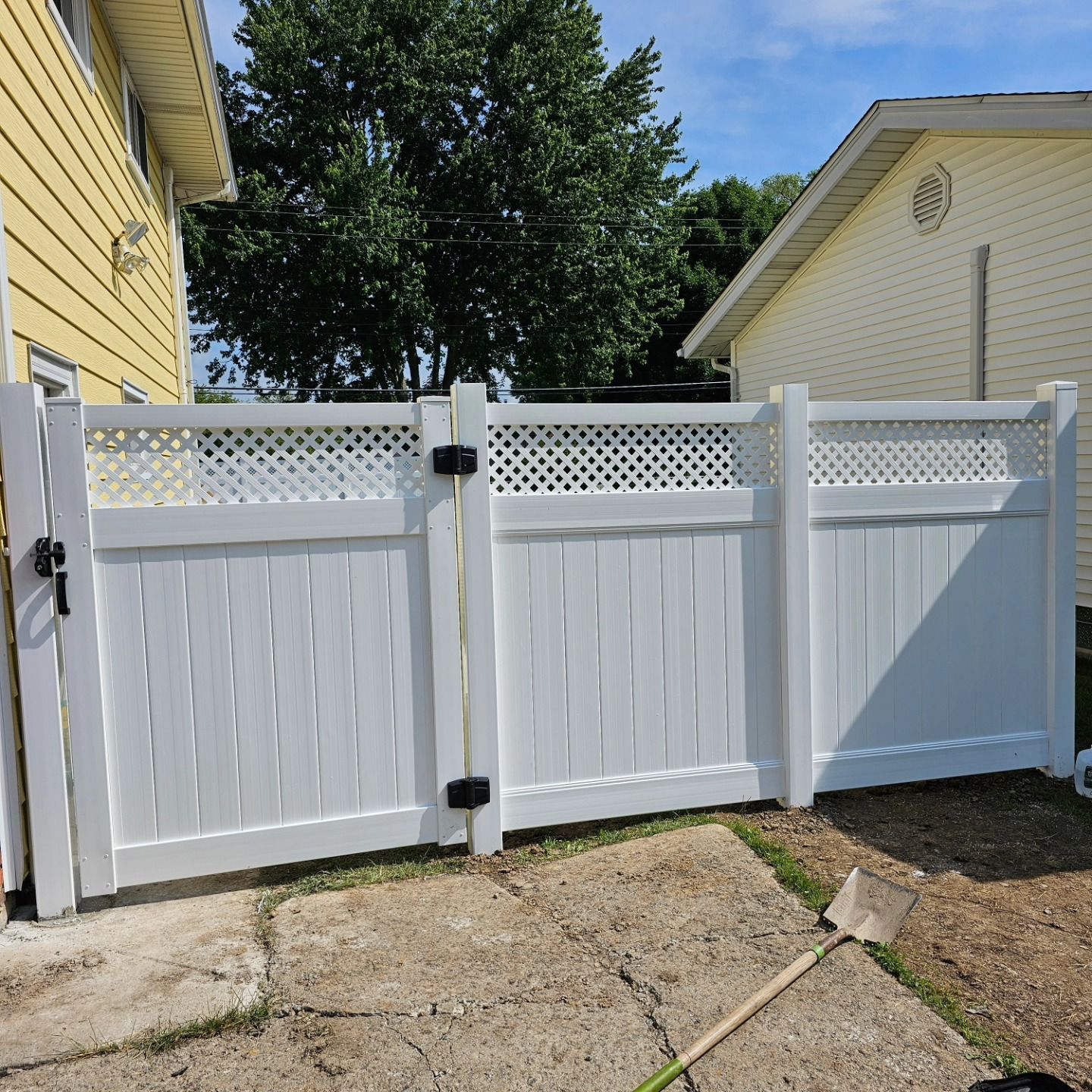 White vinyl fence gate between yellow house and white garage, with a shovel on the ground in front.