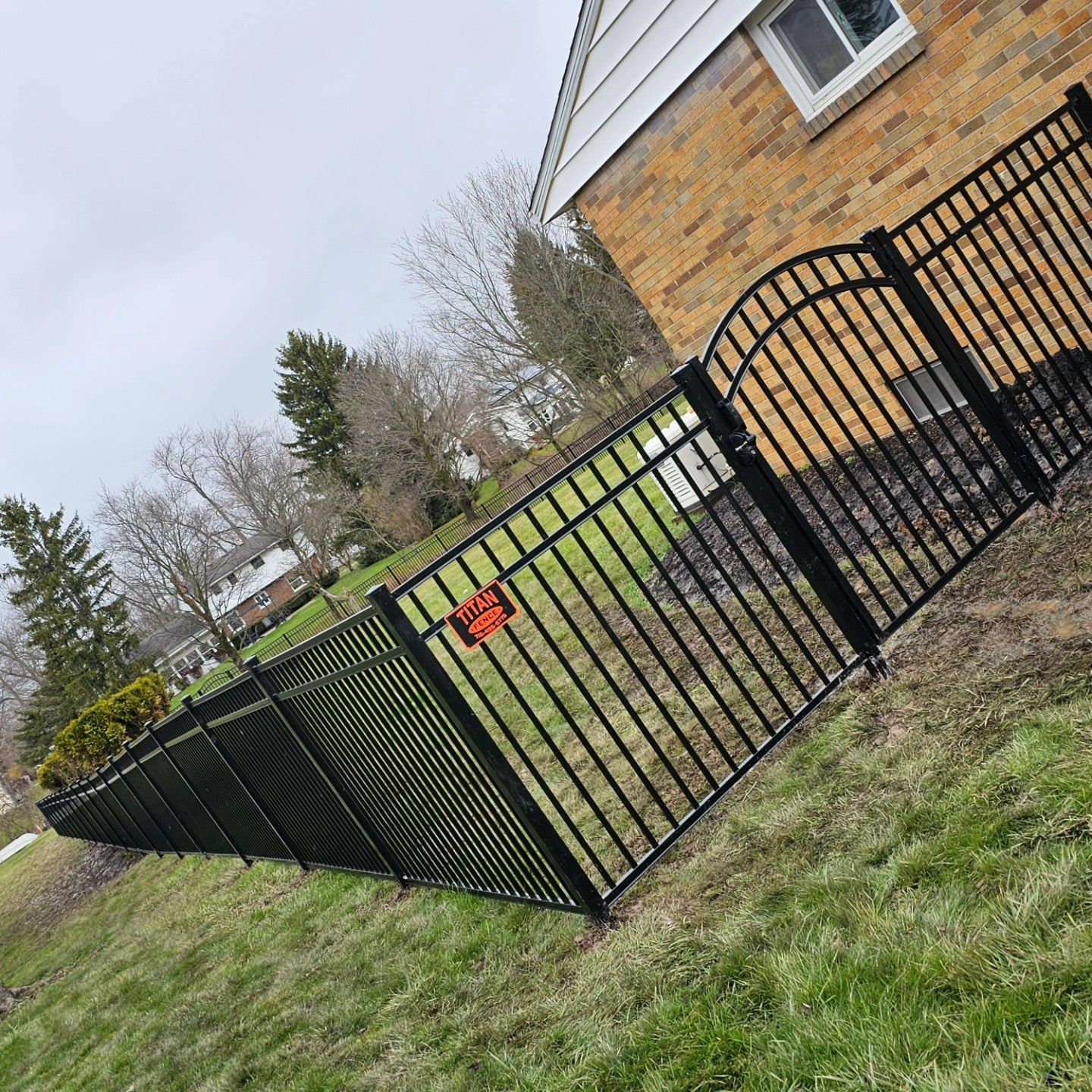 Black metal fence around a grassy yard beside a tan brick house with a red warning sign on the gate.