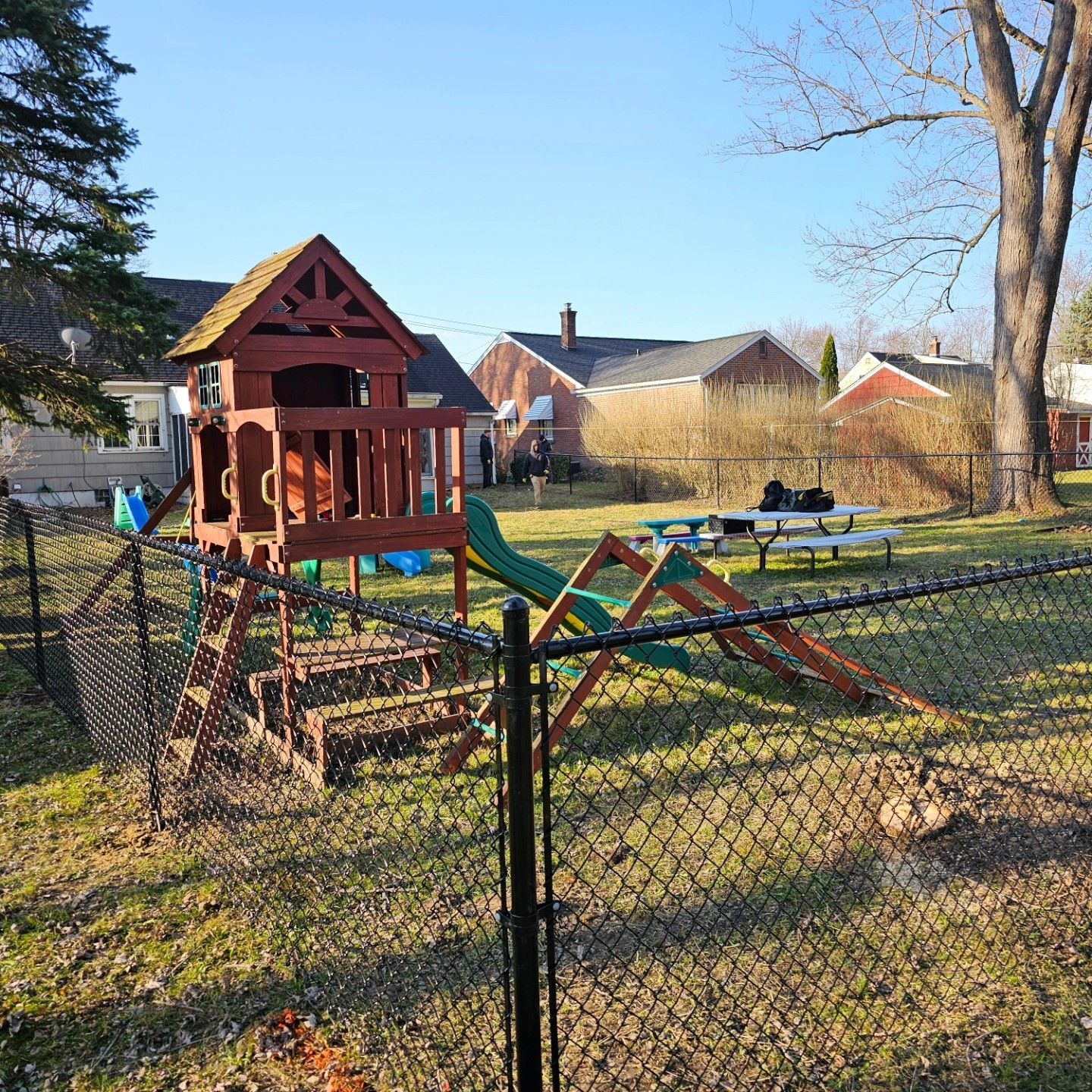 Small fenced playground with a red wooden playhouse and seesaw in a suburban yard under clear sky
