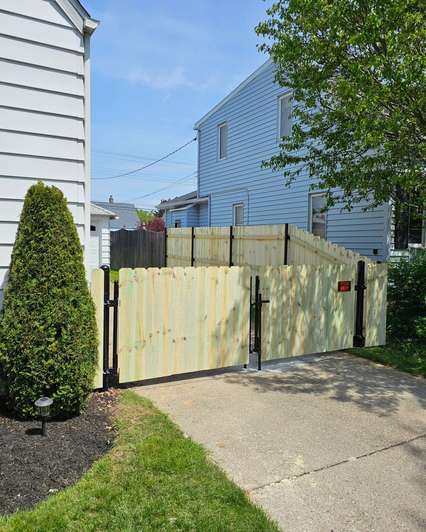Wooden driveway gate beside a sidewalk, framed by white houses and green shrubs on a sunny day