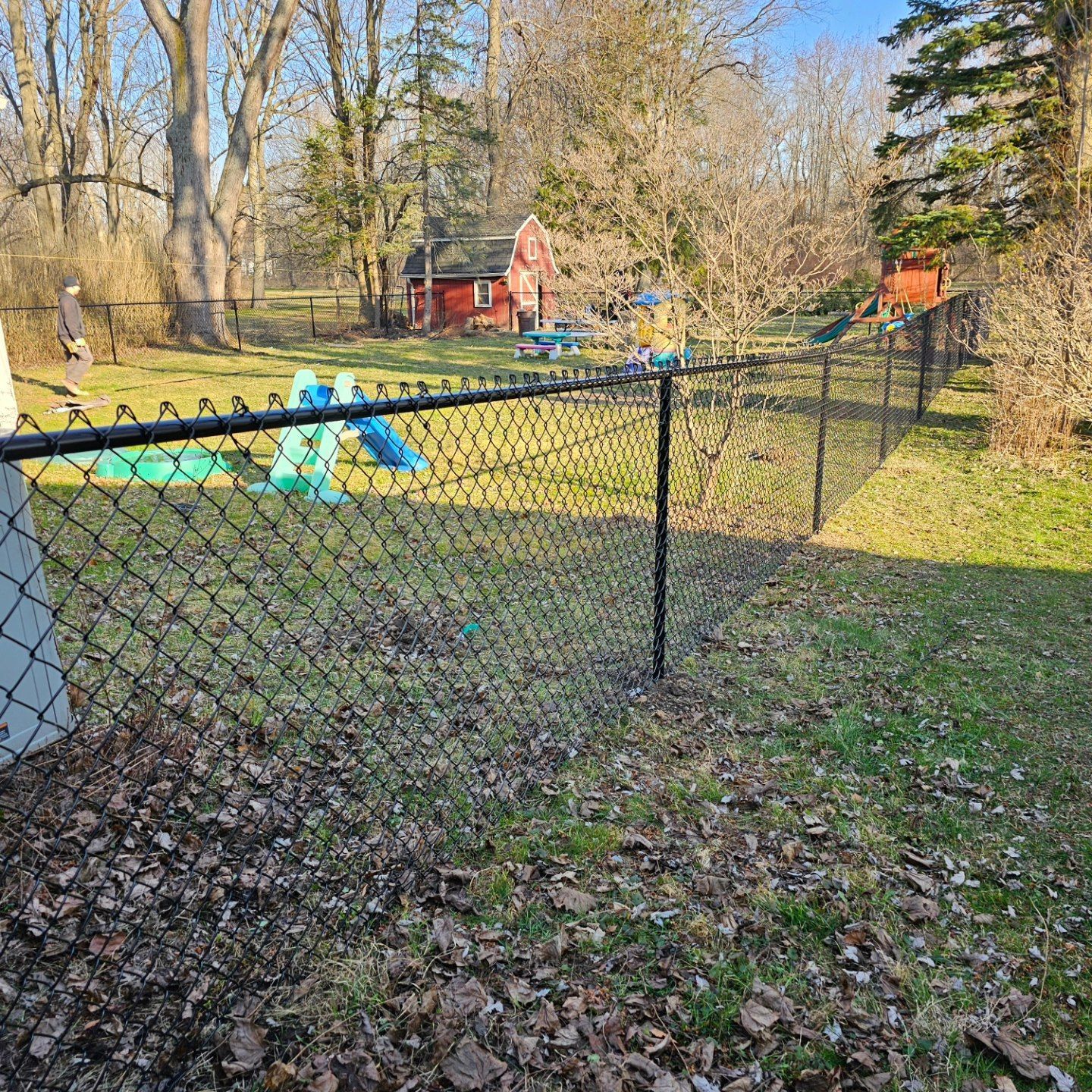 Backyard with black chain-link fence, green grass, blue play equipment, and trees in sunlight