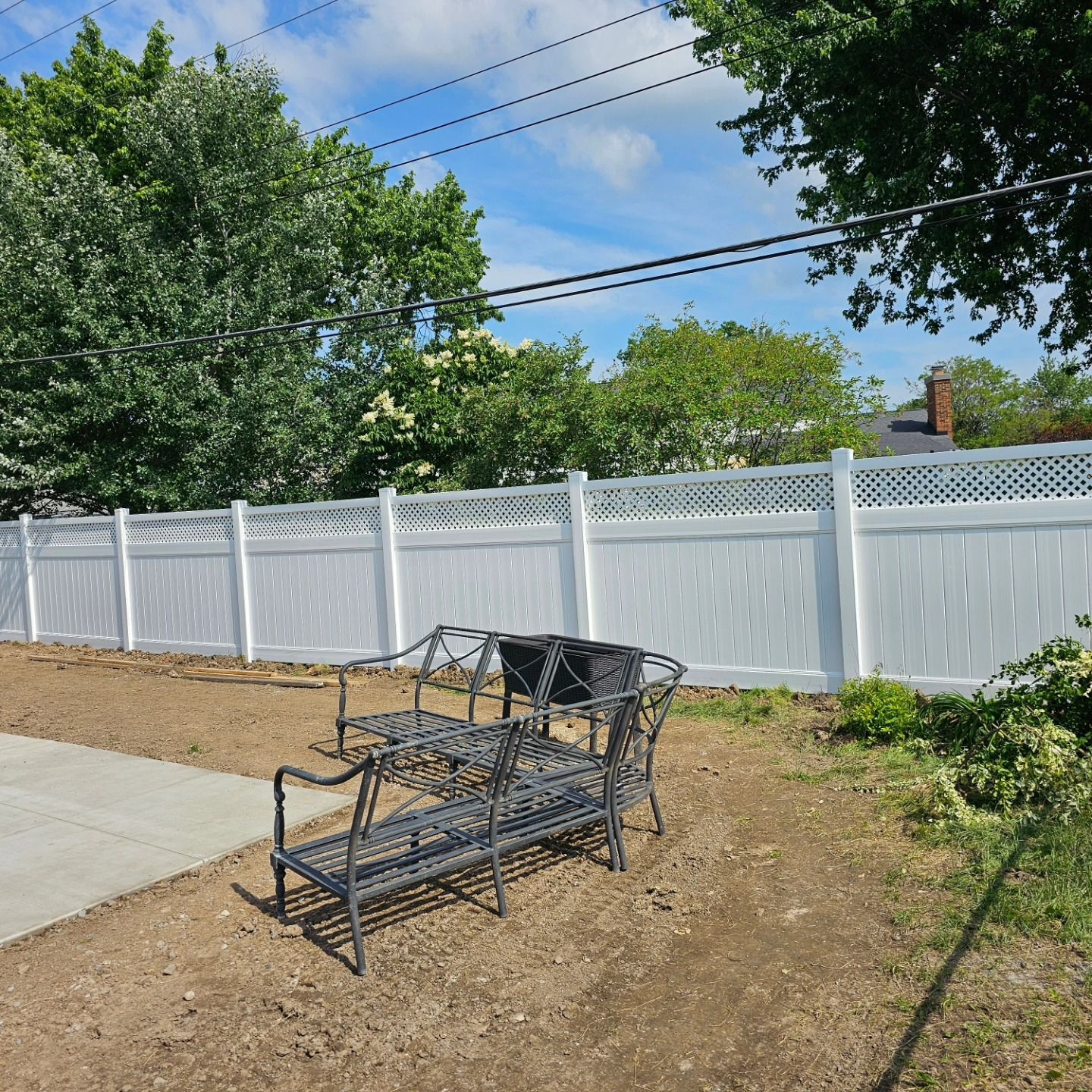 Metal patio chair and small table on a concrete patio beside a white fence and green trees under a blue sky