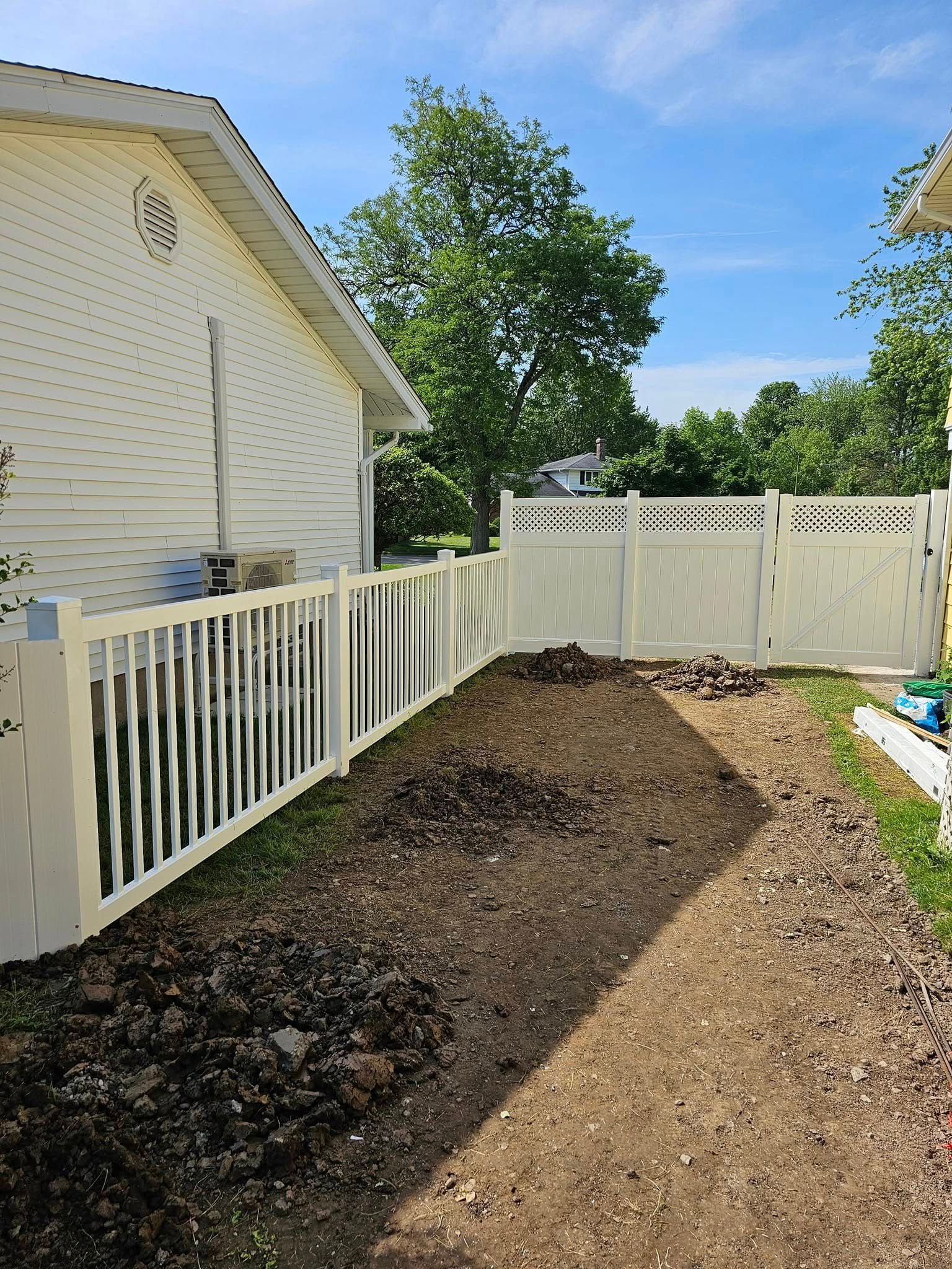 Backyard beside white house with fresh dirt pile and fenced side yard under a clear blue sky