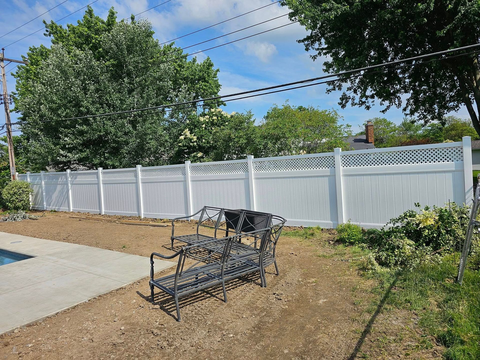 Backyard patio with a black metal loveseat, white fence, trees, and a concrete path under a blue sky