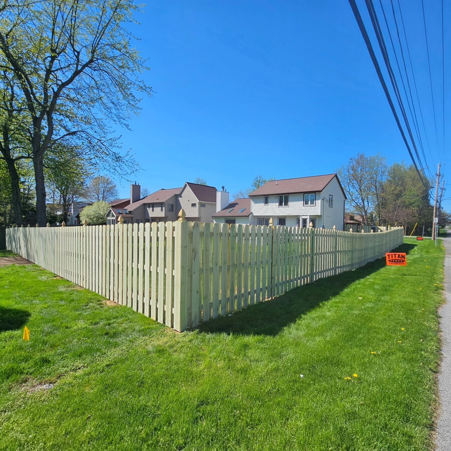Corner of a fenced suburban yard with green grass, houses, and a bright blue sky.