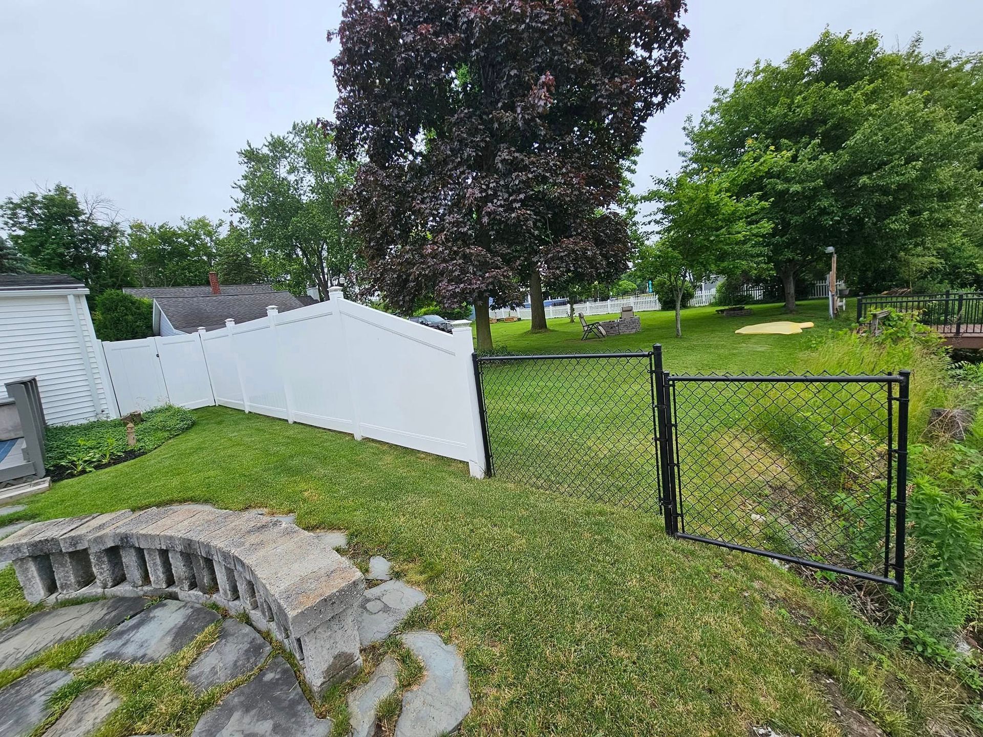 Backyard with white fence, black gate, stone steps, and large trees under an overcast sky