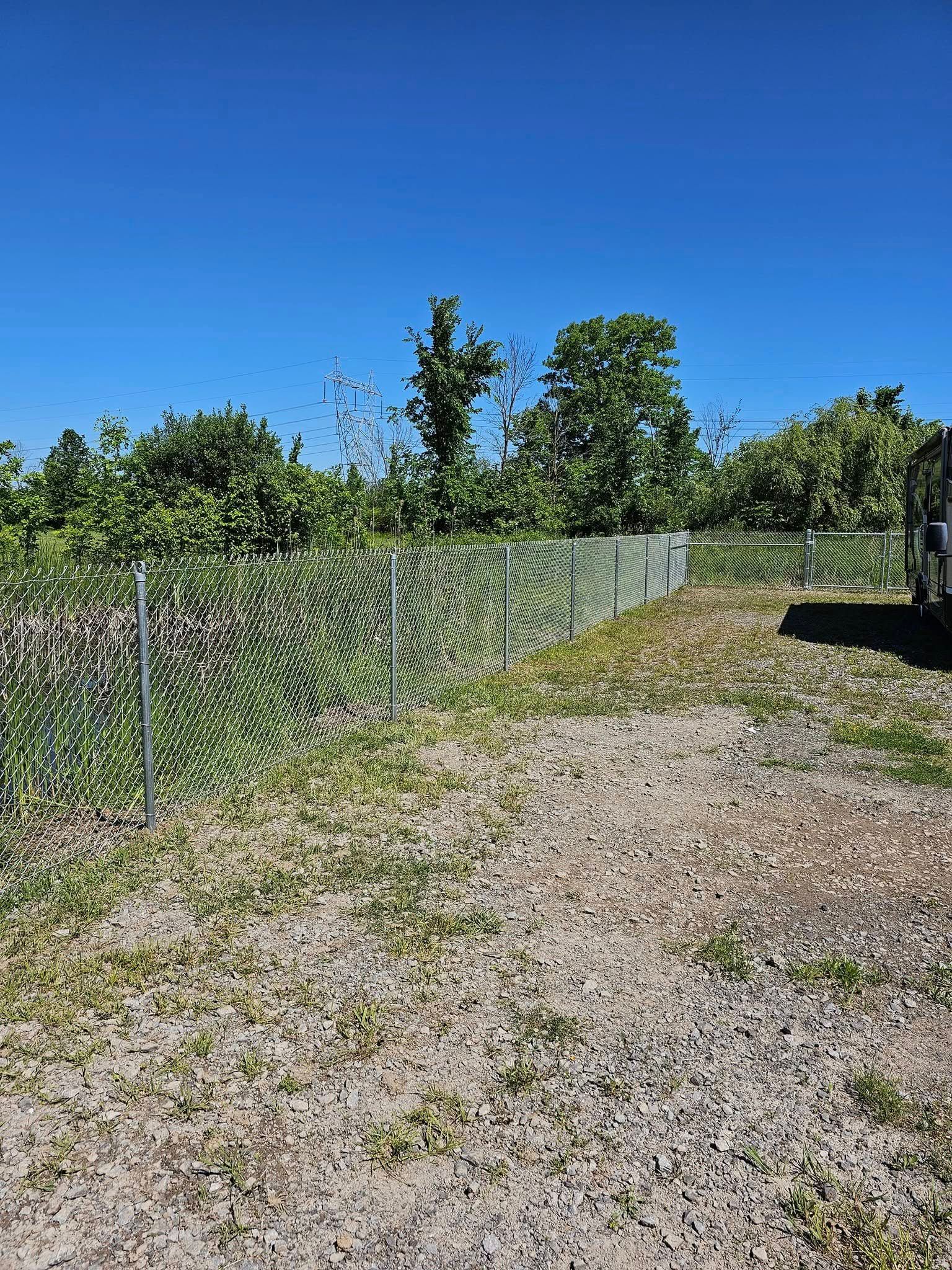 Gravel path beside a chain-link fence under a clear blue sky, with trees and a parked vehicle on the right