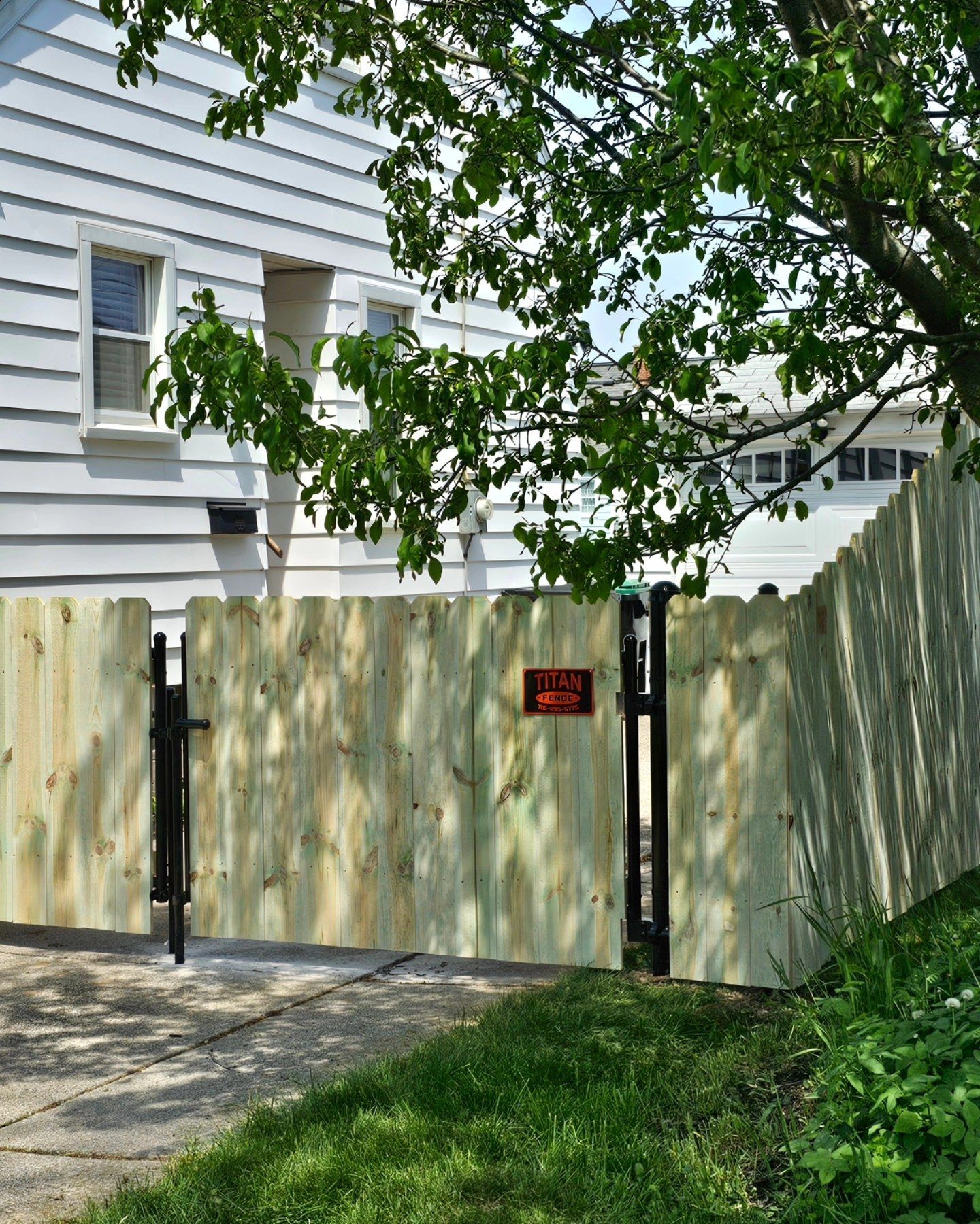 Wooden driveway gate beside a white house, with a red sign and tree branches overhead.