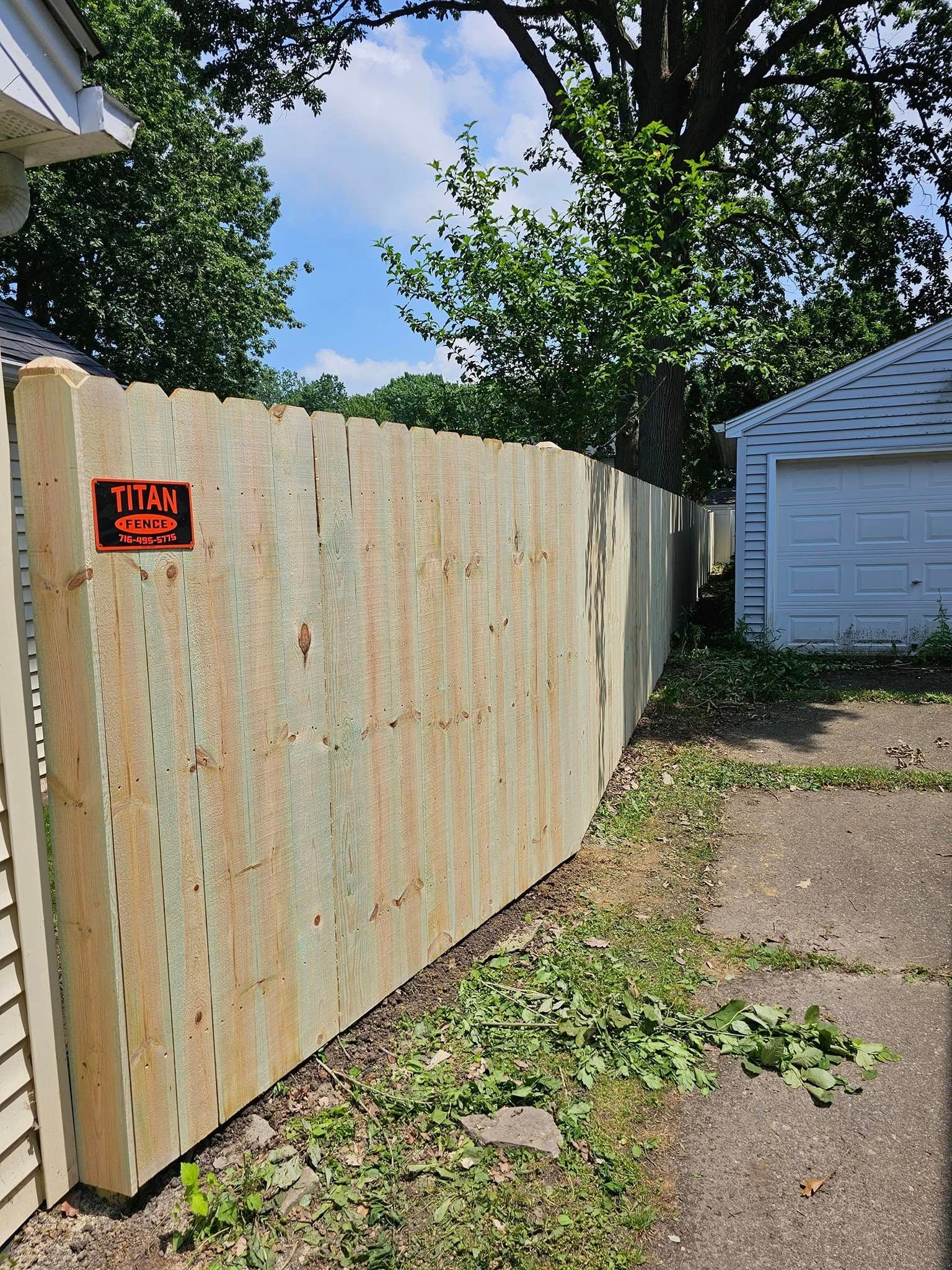 Wooden privacy fence with a red sign beside a driveway, trees overhead, and a white garage in the background