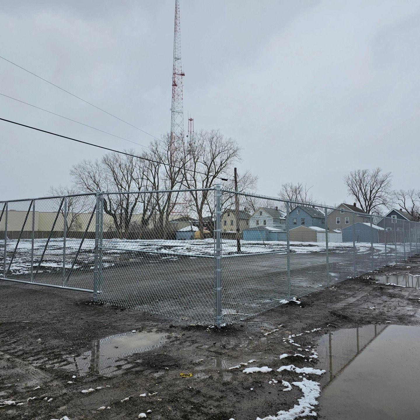 Snowy industrial lot with chain-link fence, muddy ground, bare trees, and a tall communications tower under gray sky