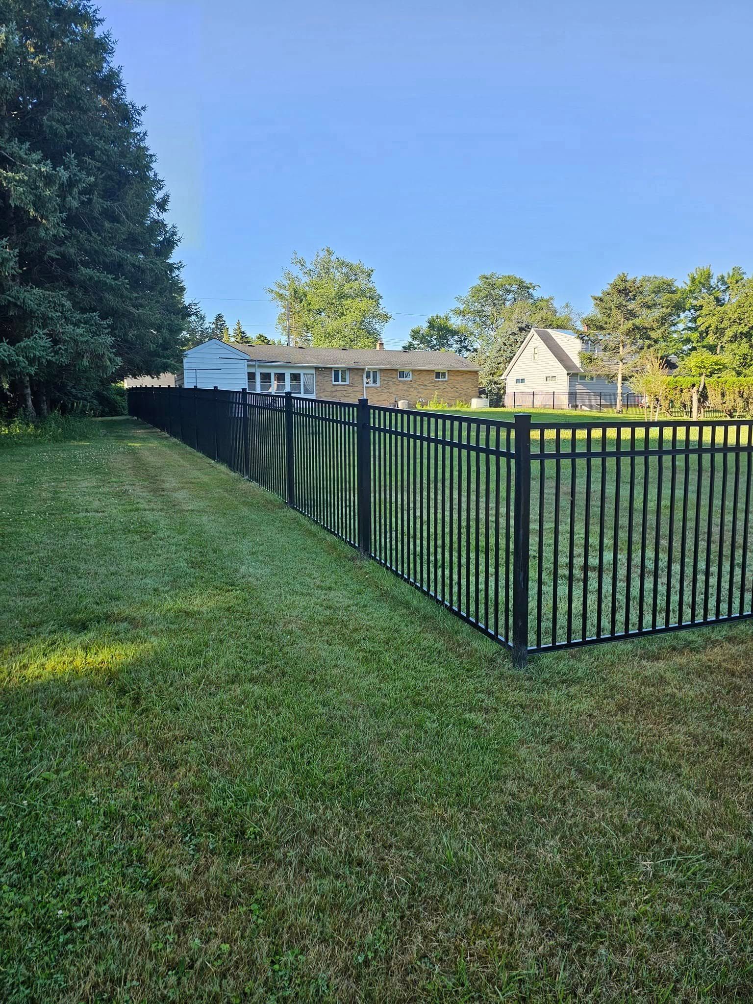 Grassy fenced backyard with a black metal fence, tall trees, and houses in the distance.